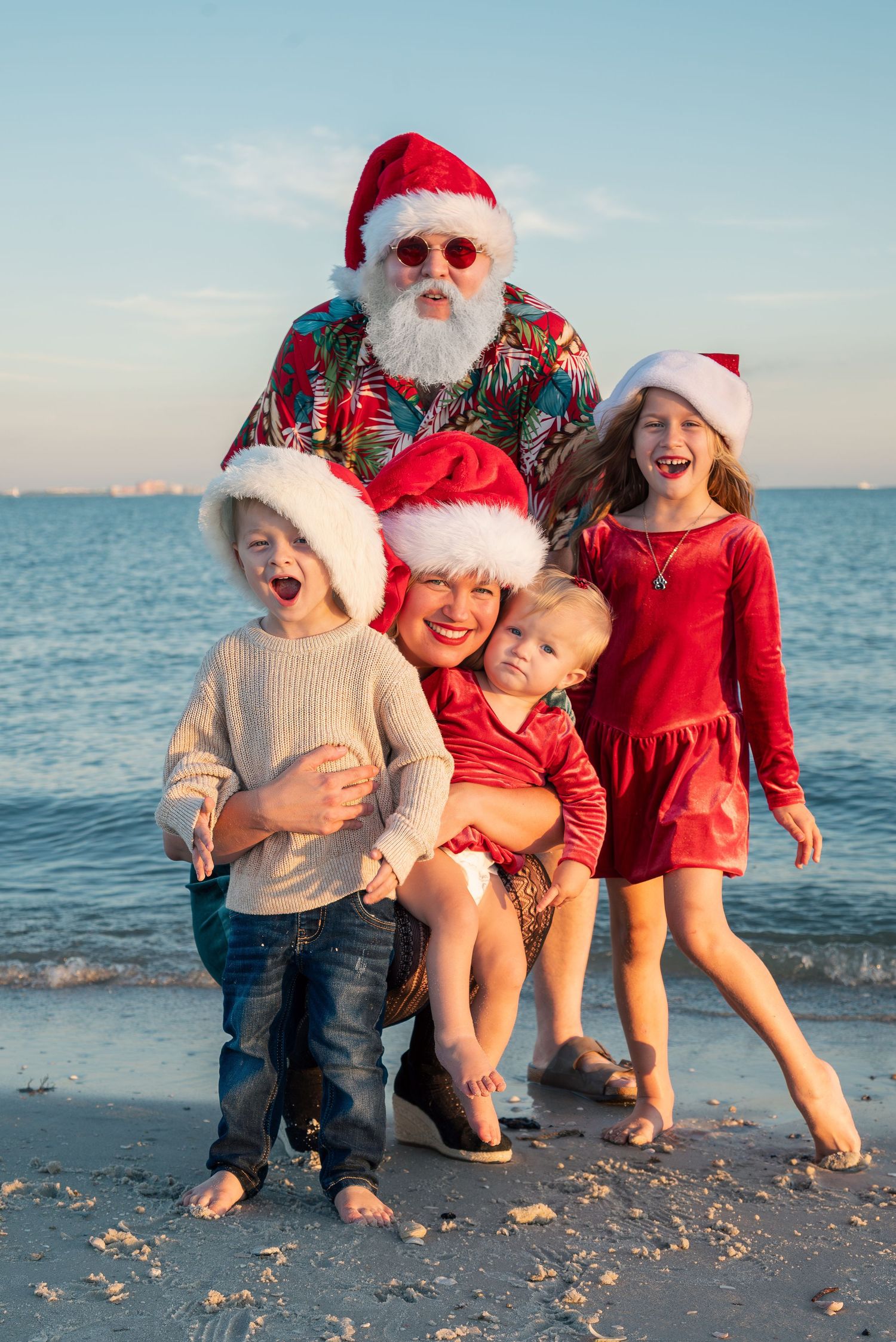 Family with beach Santa Claus and three children laughing together on a sunny shoreline.