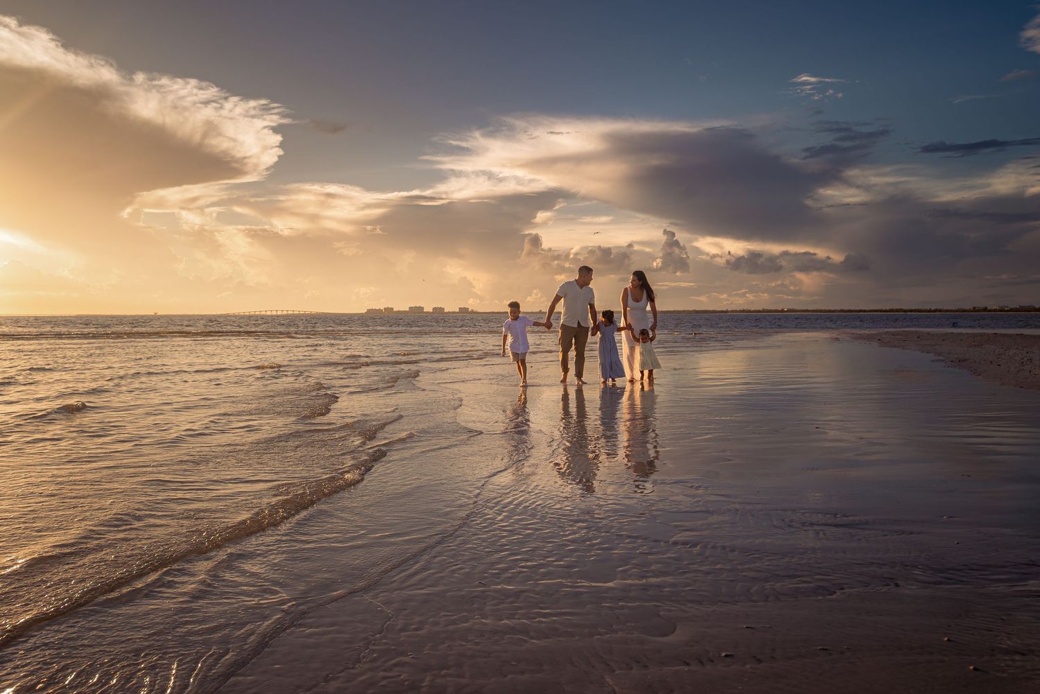 A family of four walks hand-in-hand along a reflective beach at golden sunset with dramatic cloudy skies.