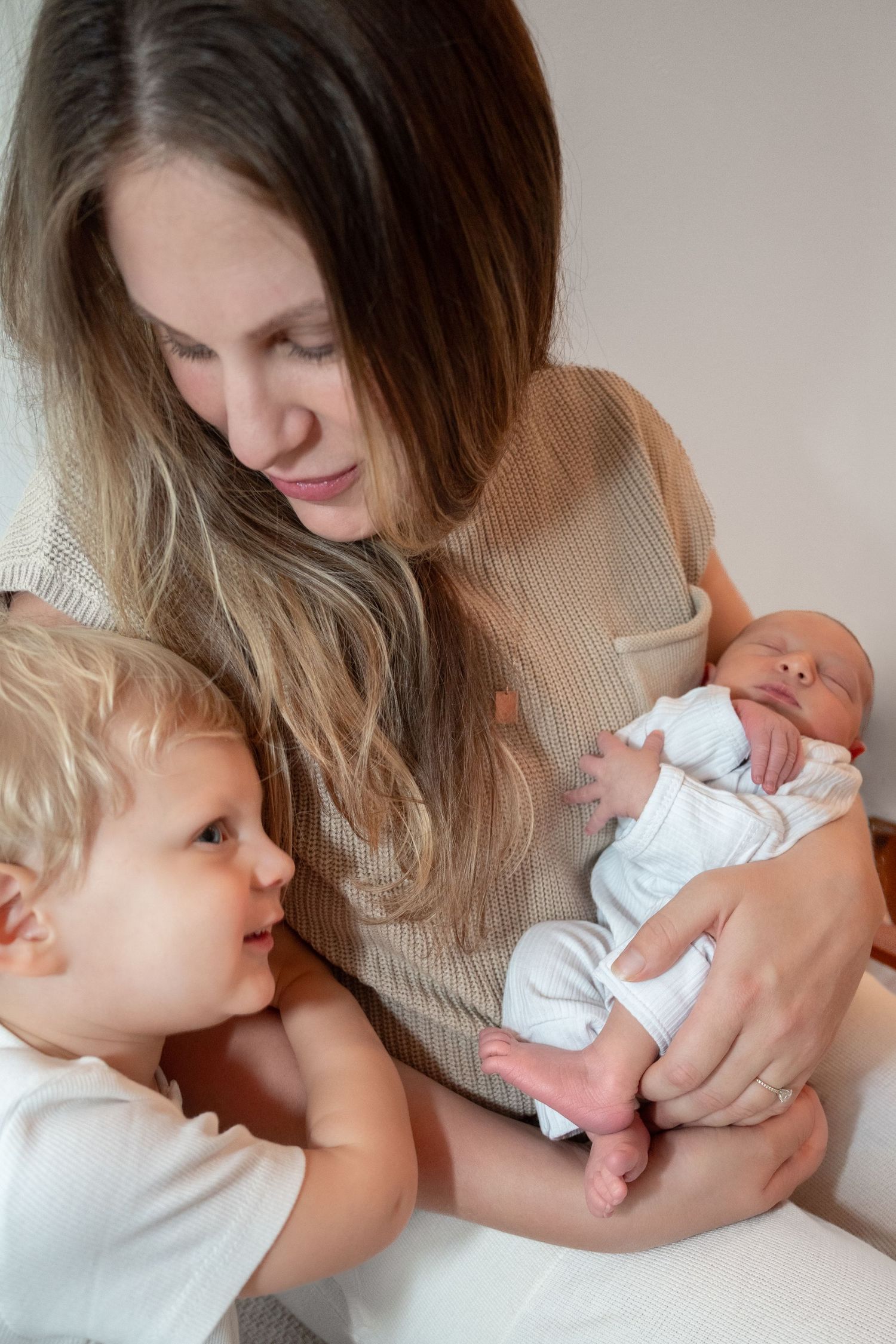 Mother holds newborn baby while toddler sibling looks on with curiosity indoors.