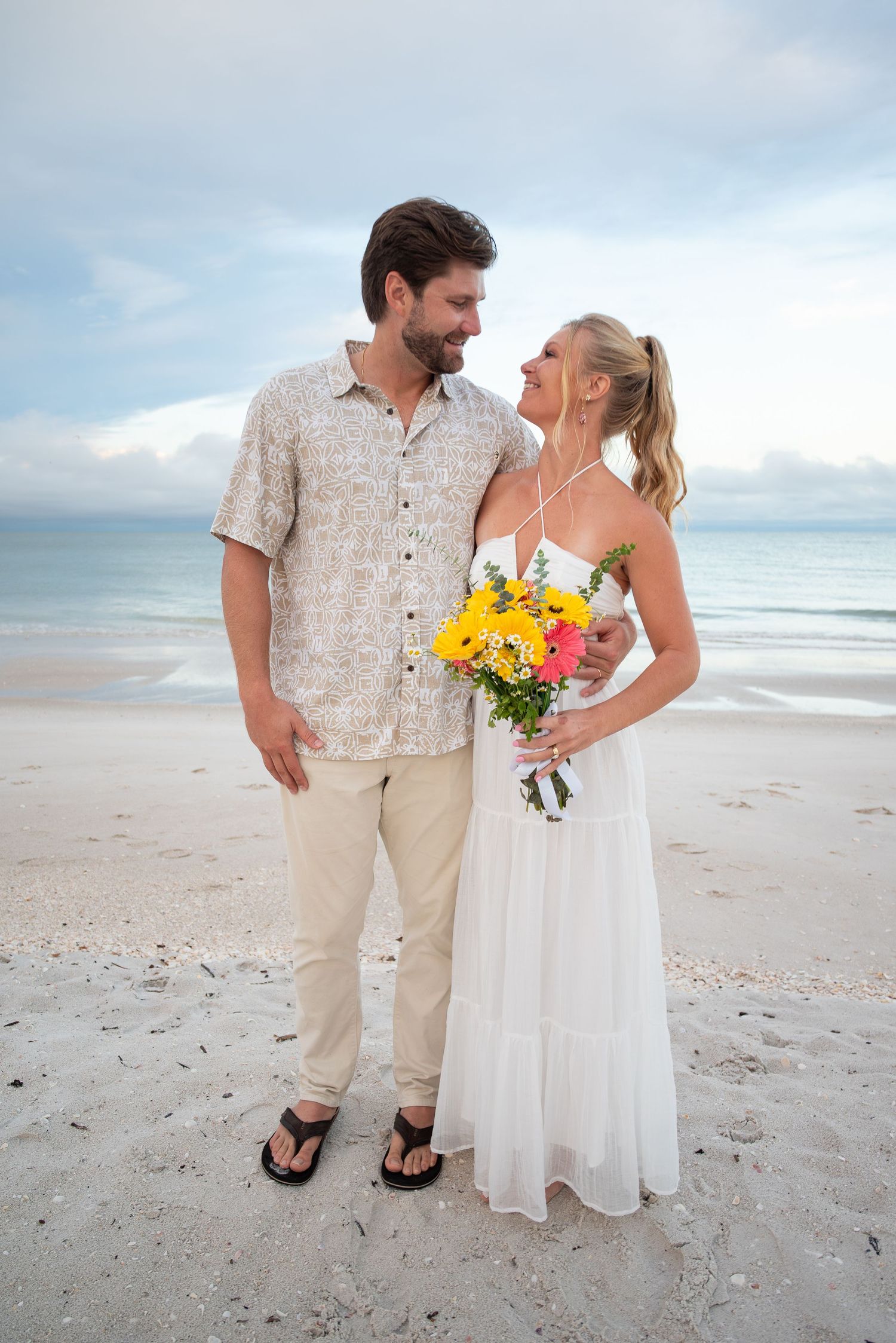 Couple in beach wedding attire, woman in white dress holding colorful bouquet, man in floral shirt, ocean backdrop.