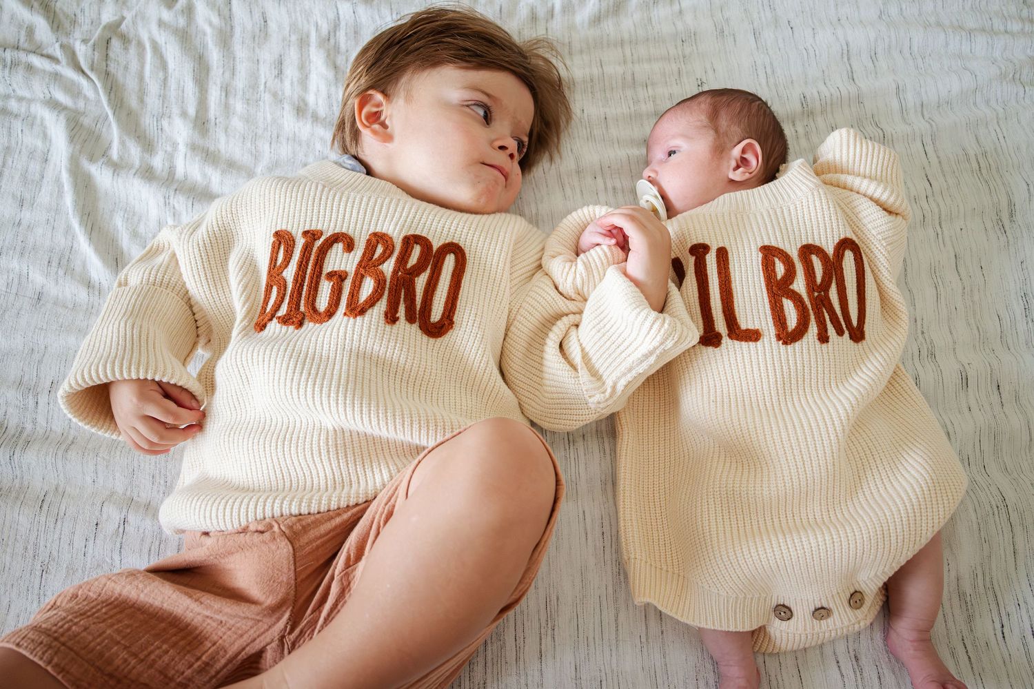 Two brothers in matching cream knit sweaters labeled Big Bro and Lil Bro lying on a white bed.