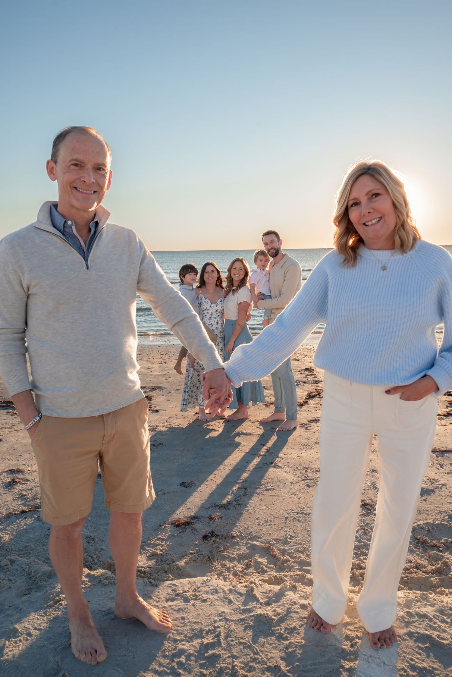 Smiling couple holding hands on beach with family group behind them.