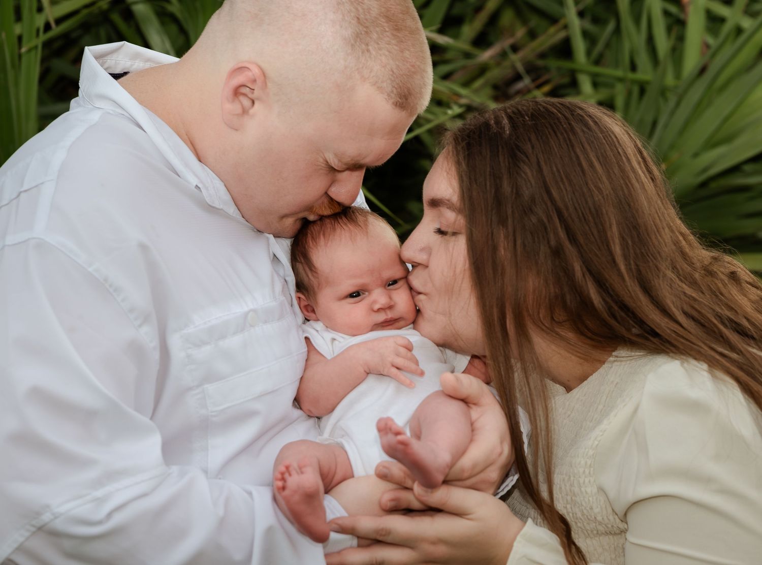 Happy new parents kissing their newborn baby together in a loving outdoor portrait.