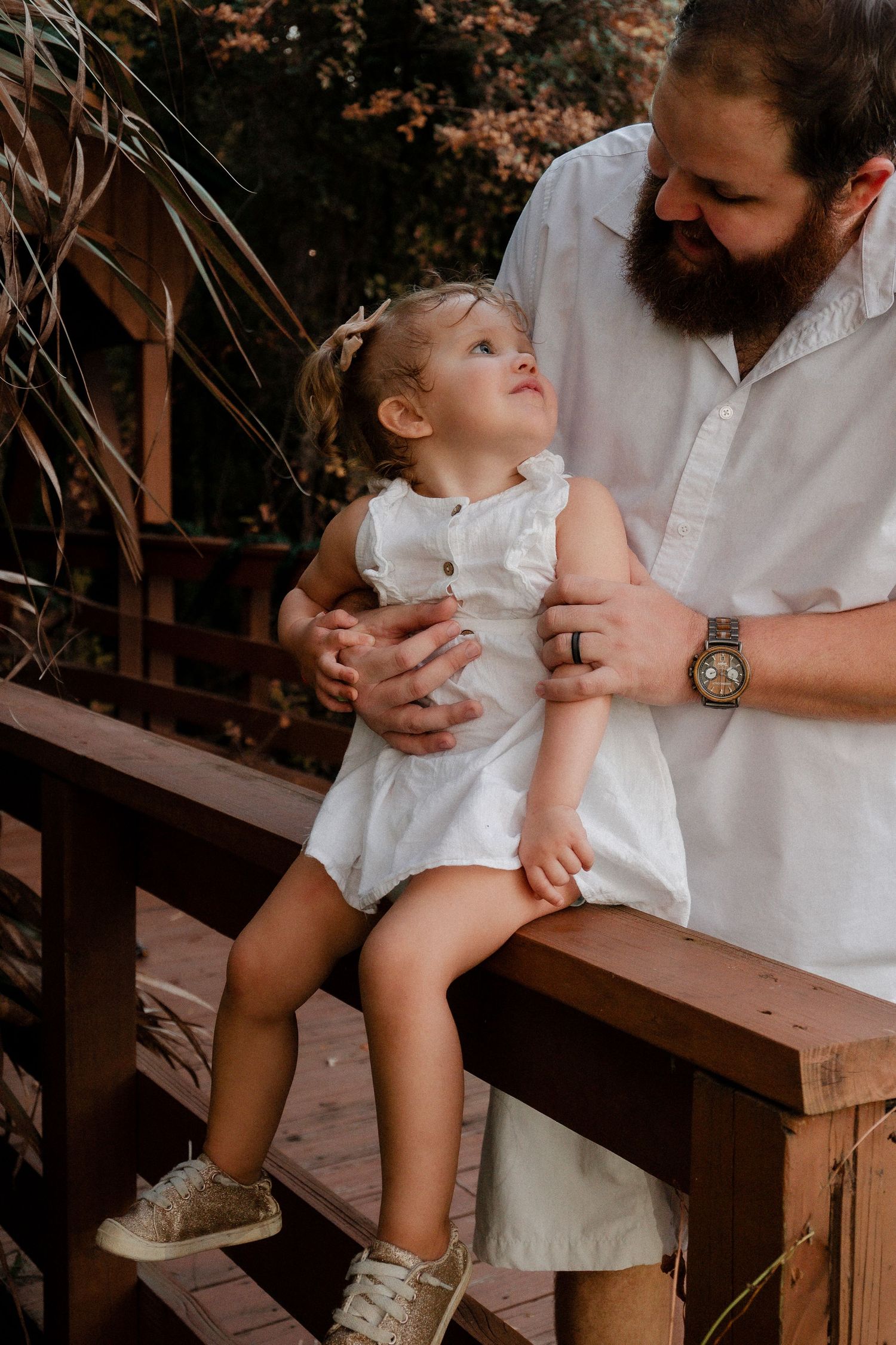 A bearded father holds his toddler daughter on a wooden railing surrounded by nature.