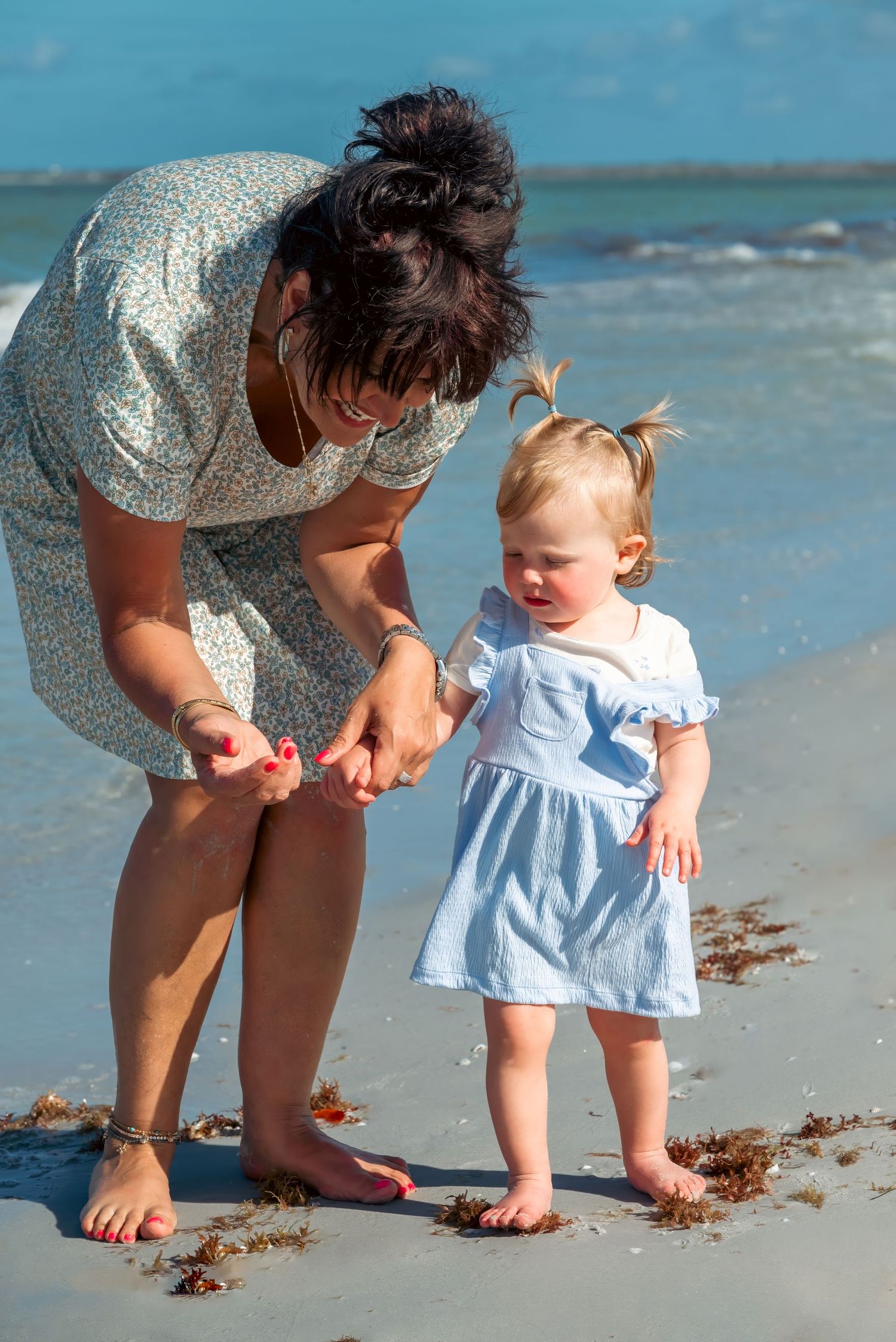 Mother and toddler girl in blue dress playing on sandy beach near ocean waves on a sunny day.