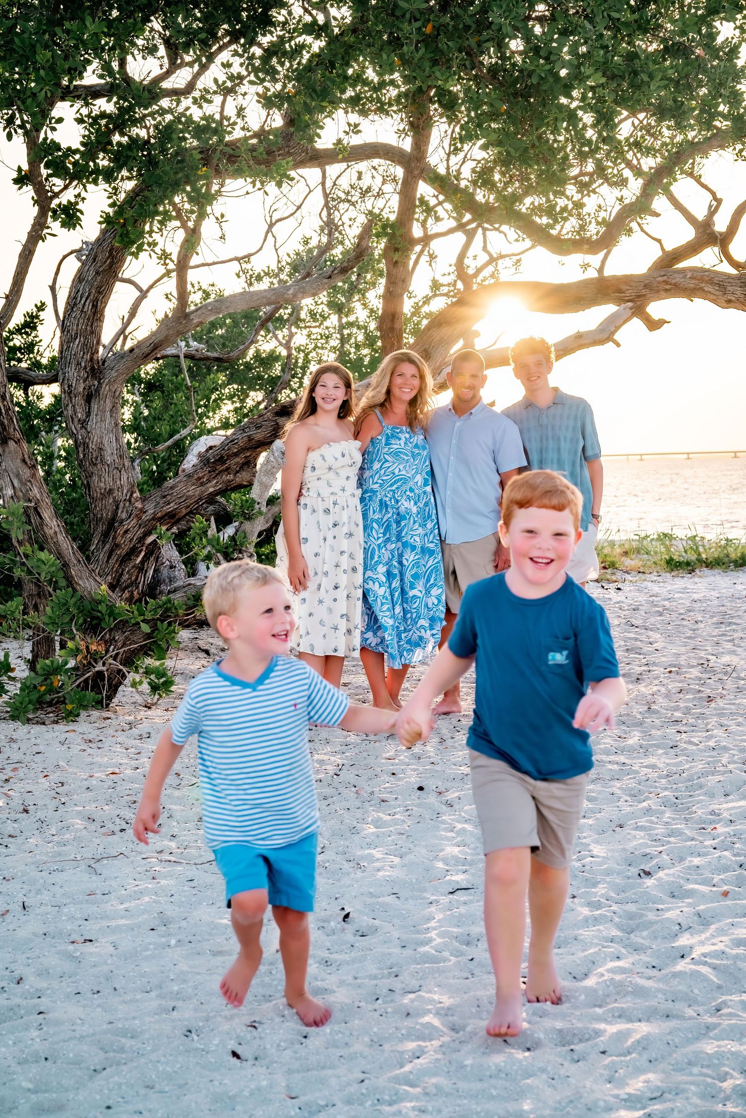 Family of five enjoying a golden sunset beach walk, two young red-haired boys running on white sand near a tropical tree.