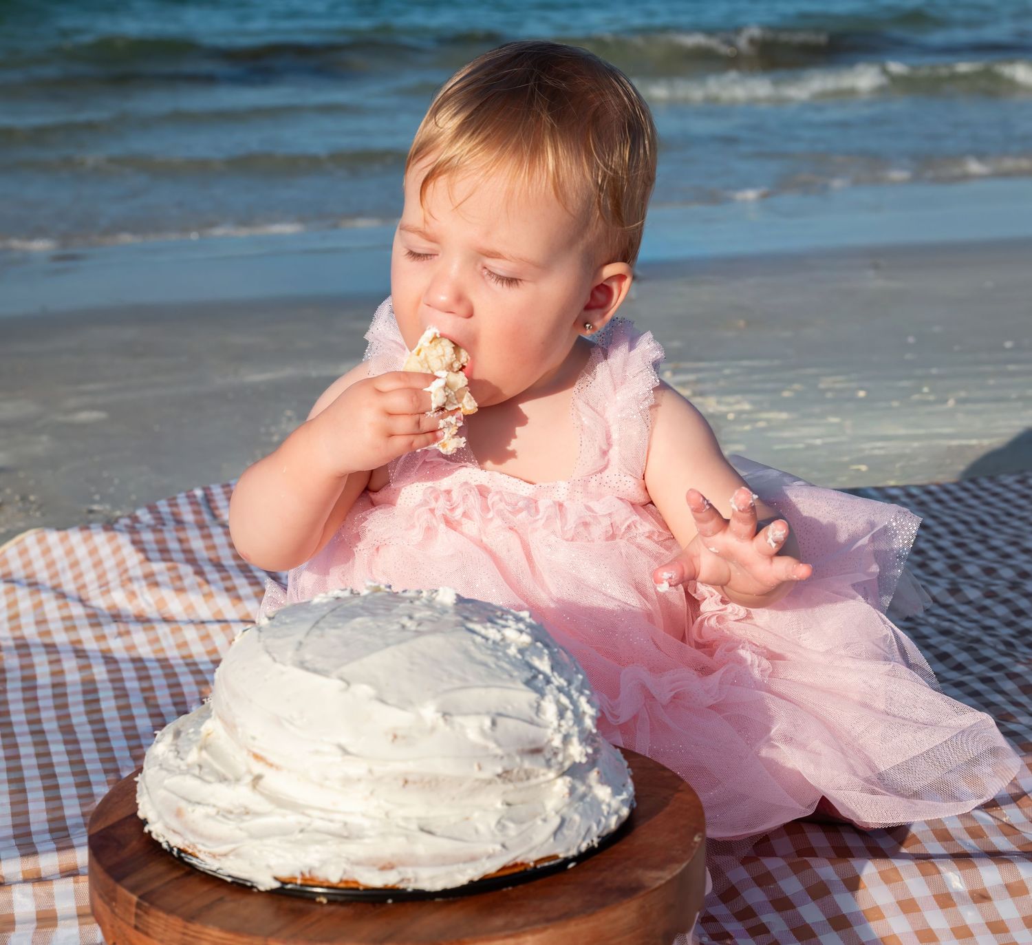 Adorable baby in pink ruffle dress eating smash cake on beach blanket during first birthday celebration.