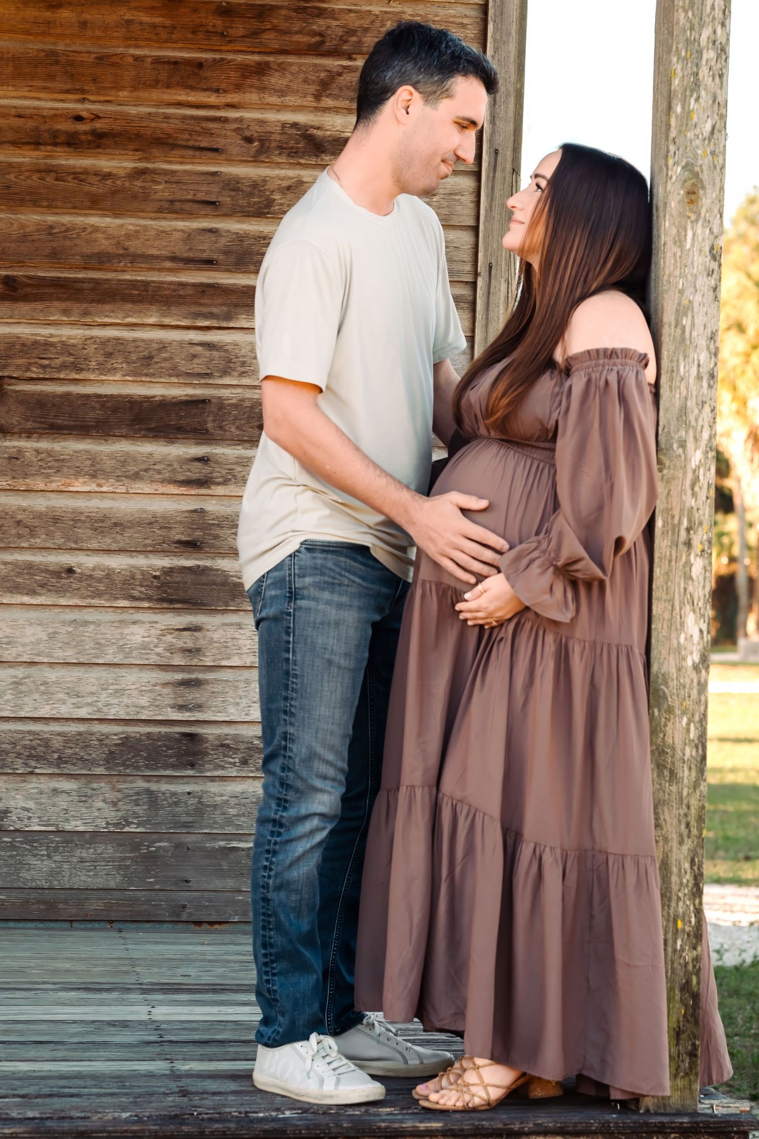 Expecting couple sharing a tender moment on wooden porch steps beside a rustic log cabin wall.