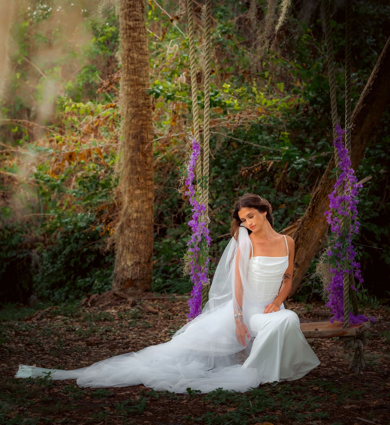 Beautiful bride in white gown sits gracefully on a wooden swing surrounded by purple flowers in a lush garden.