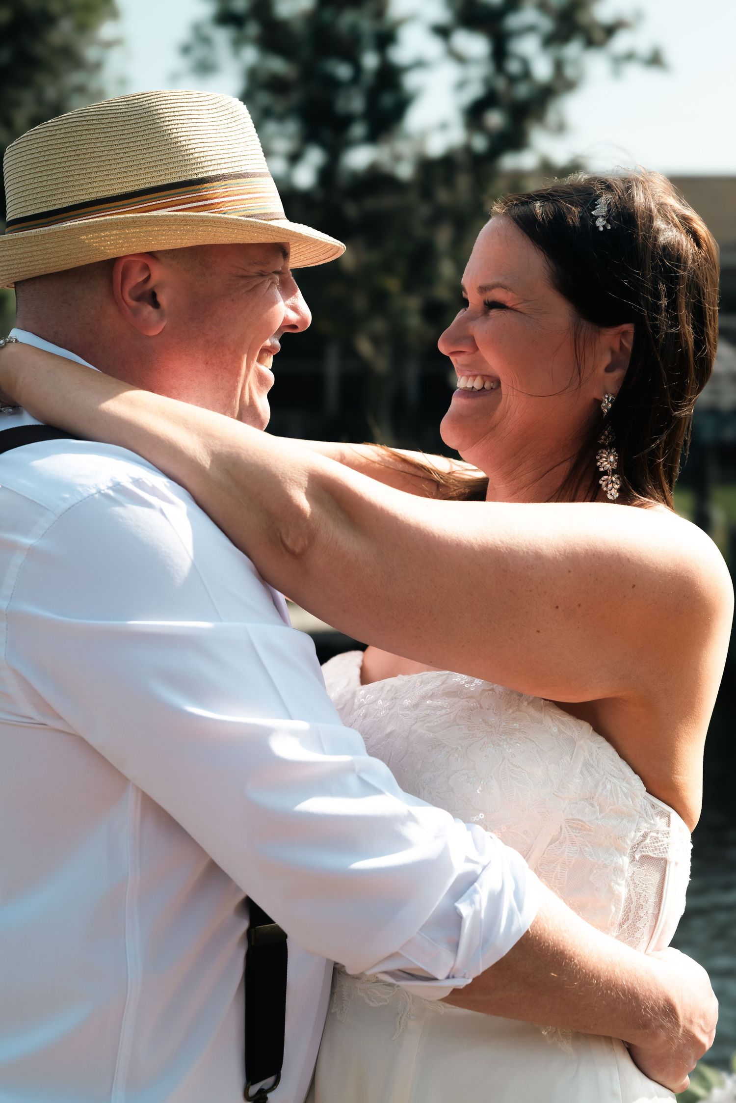 Joyful bride and groom embracing and laughing together outdoors on their wedding day, groom wearing a straw fedora hat.