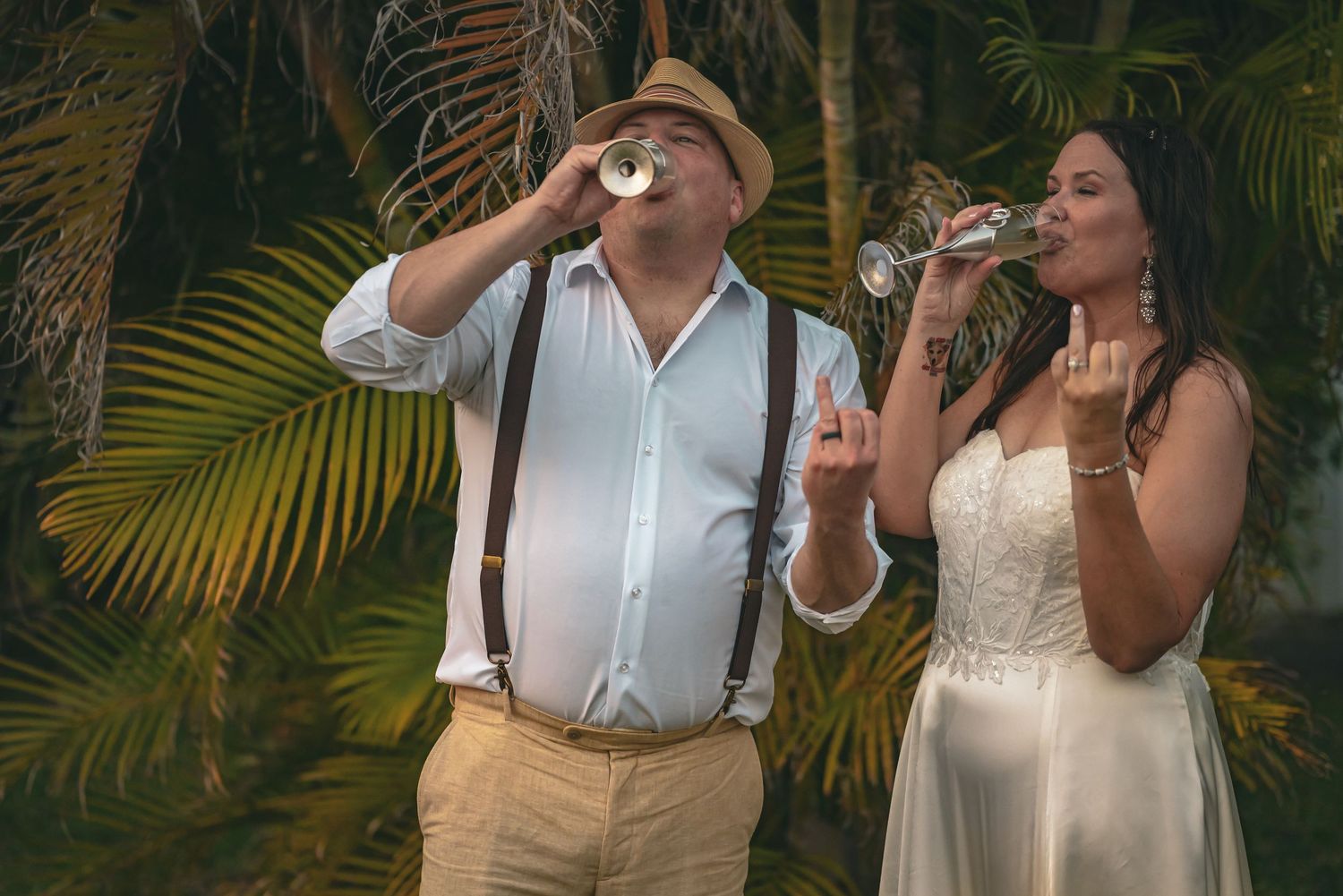 Bride and groom toasting with drinks surrounded by tropical palm trees at outdoor wedding.