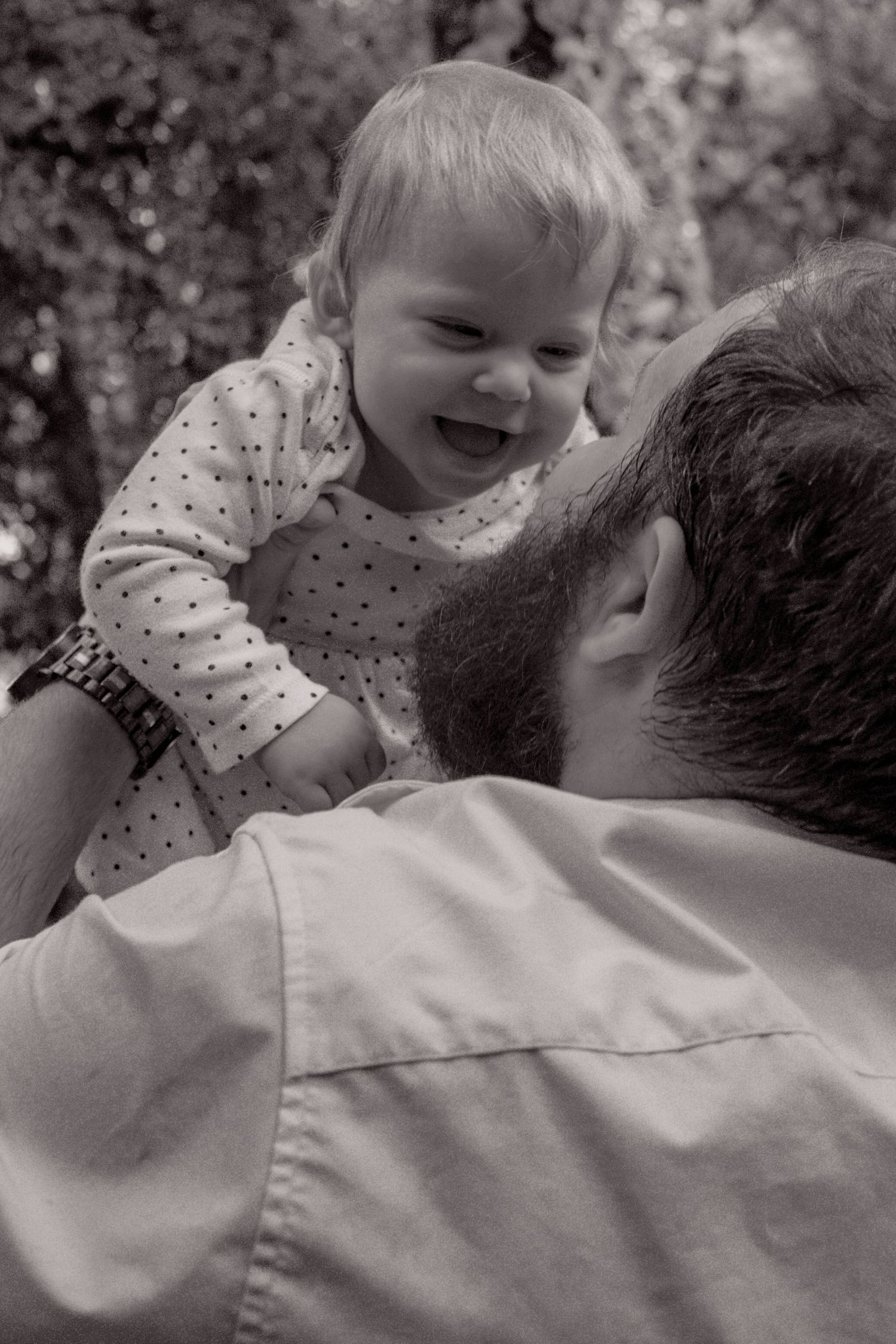Black and white photo of laughing baby being lifted by father in outdoor setting.