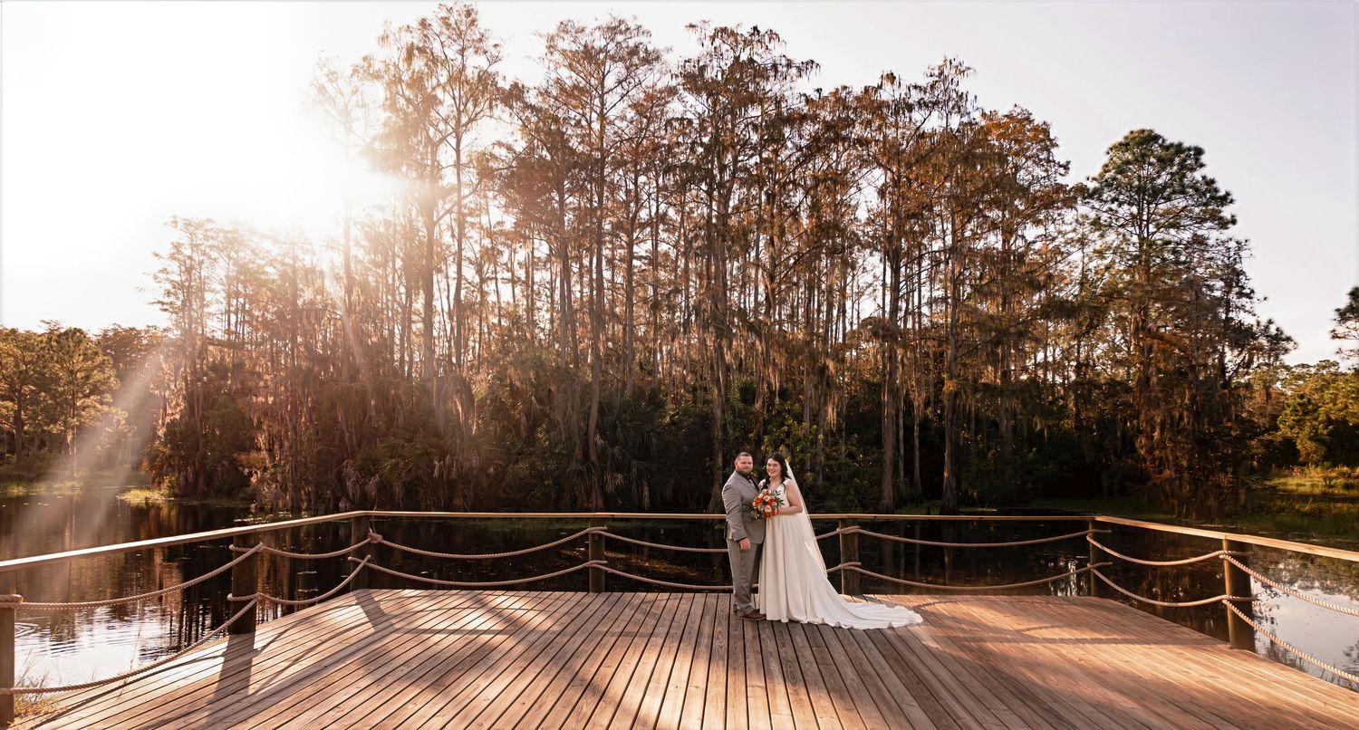 Bride and groom pose on a wooden deck overlooking a serene lake with sunlight streaming through trees.
