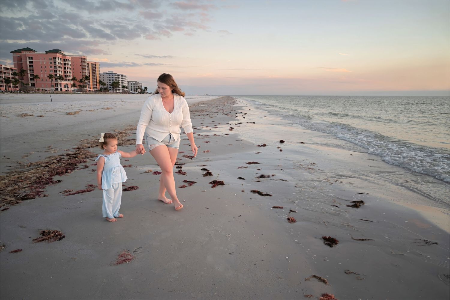Mother and young daughter in white outfits walking hand in hand along a serene beach at sunset.