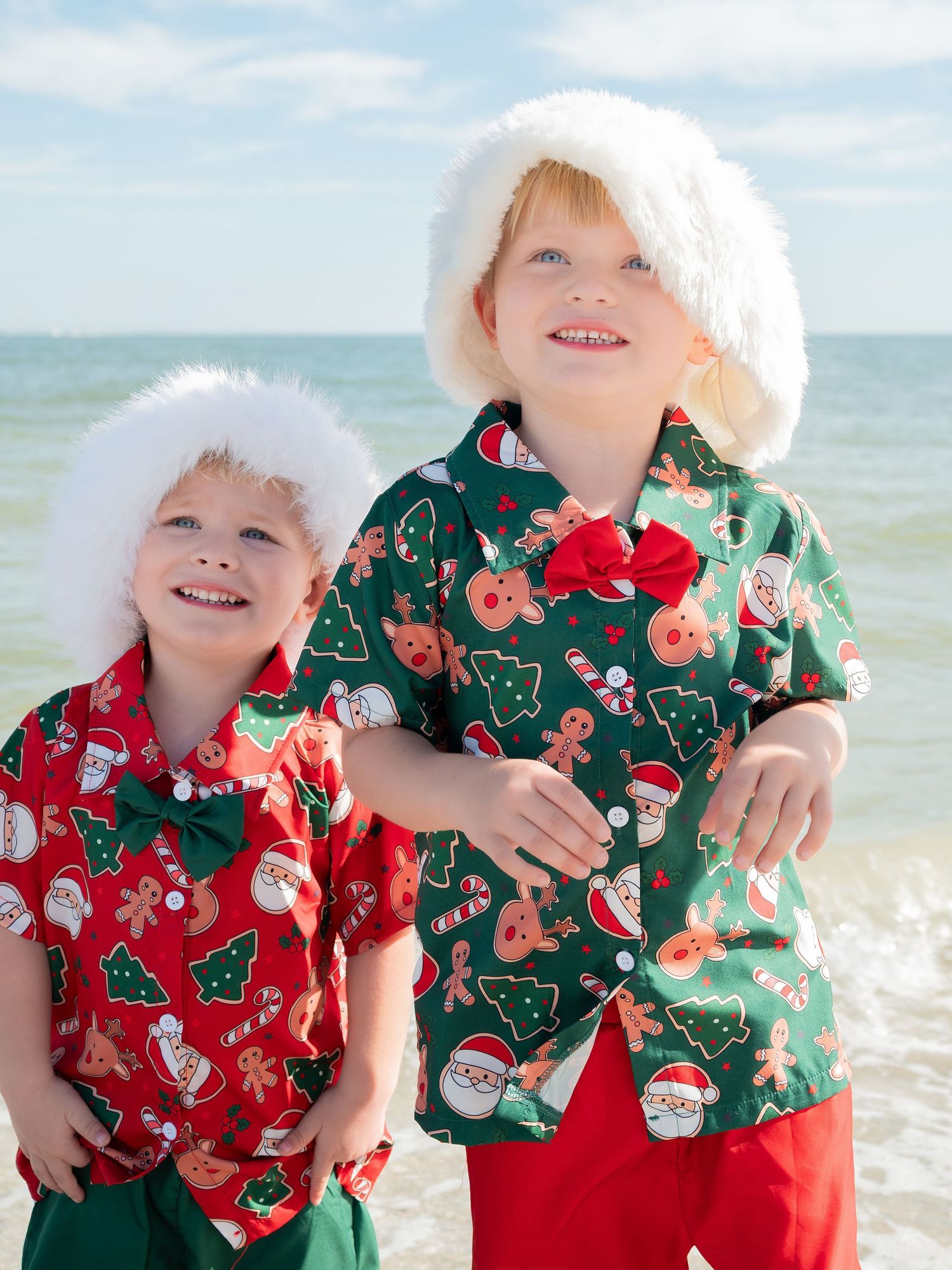 Two young boys in Christmas shirts and Santa hats smiling on a sunny beach.