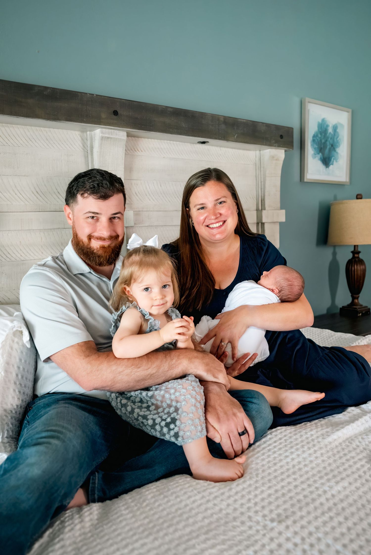 Happy family of four poses on bed, parents with toddler girl and newborn baby, smiling in cozy blue bedroom.