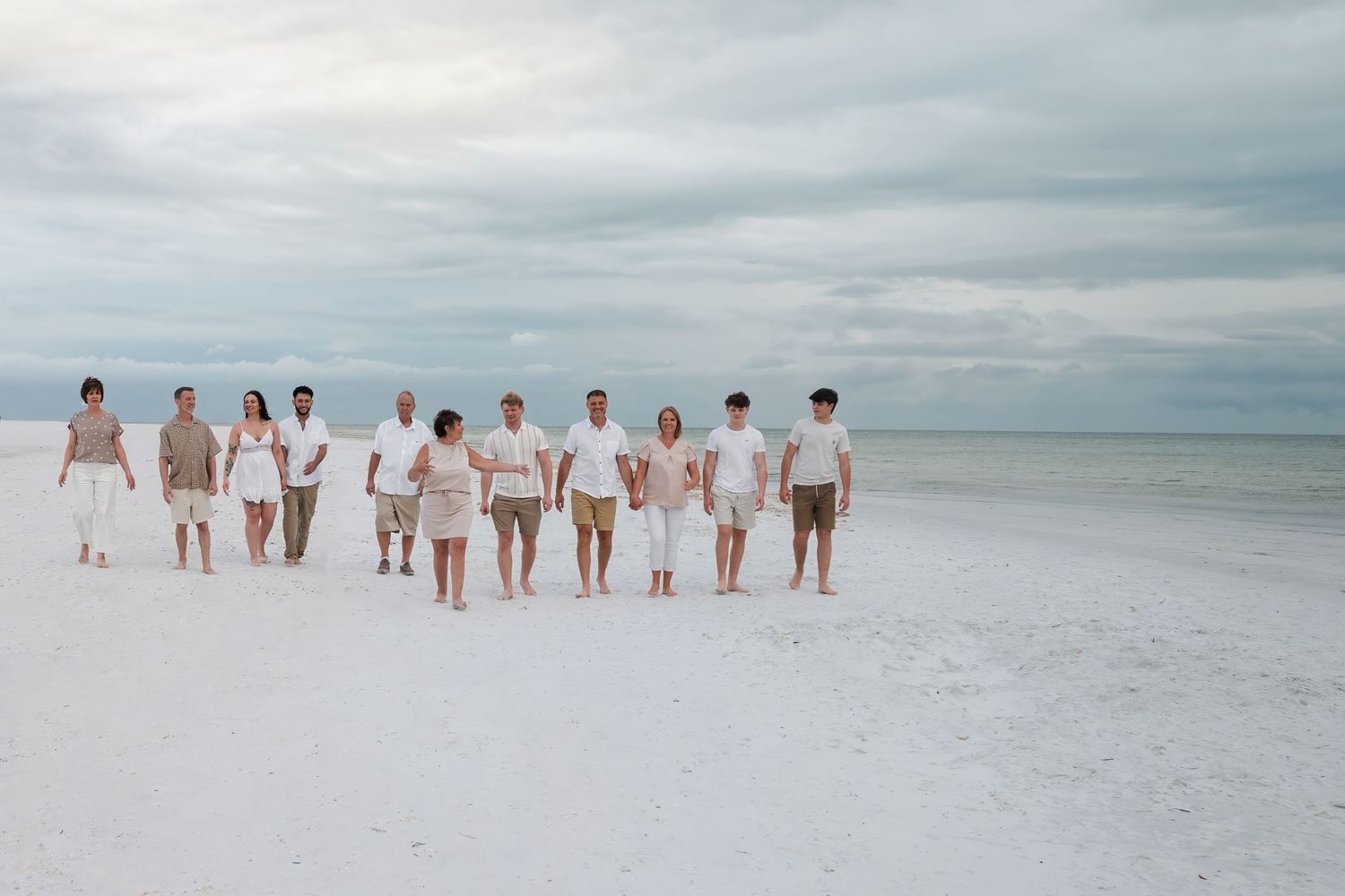 Large family group walking together on a white sandy beach under cloudy skies wearing coordinated white and tan outfits.