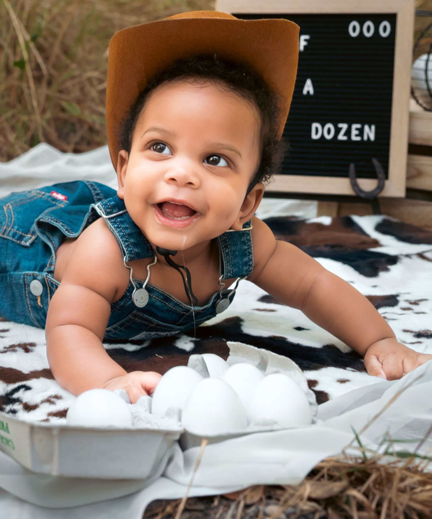 Smiling baby in cowboy hat and denim overalls lying on blanket with eggs and letter board sign outdoors.