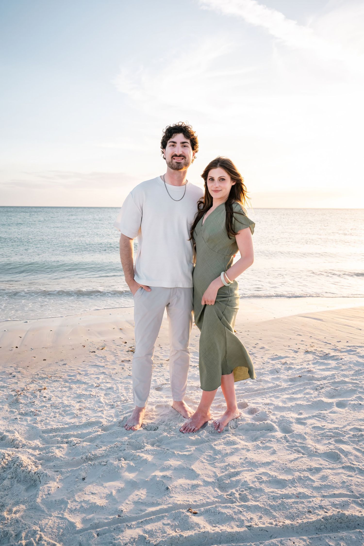 Couple standing barefoot on a sandy beach at sunset, man in white shirt and woman in green dress.