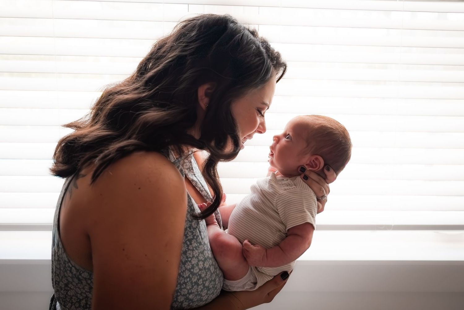 A mother tenderly holds her newborn baby, gazing lovingly as soft light filters through window blinds.