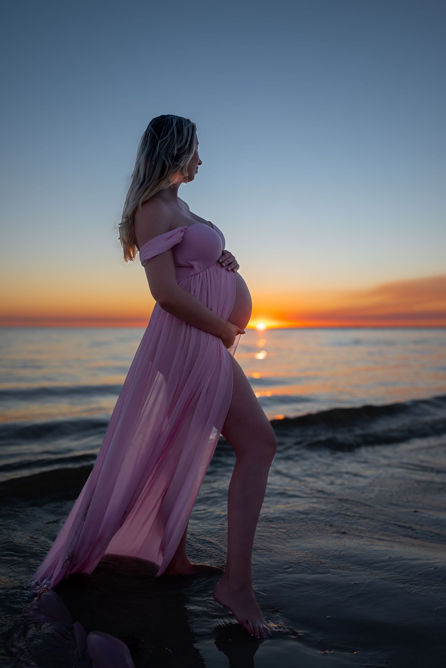 Pregnant woman in a flowing pink gown standing on the beach at a vibrant sunset.