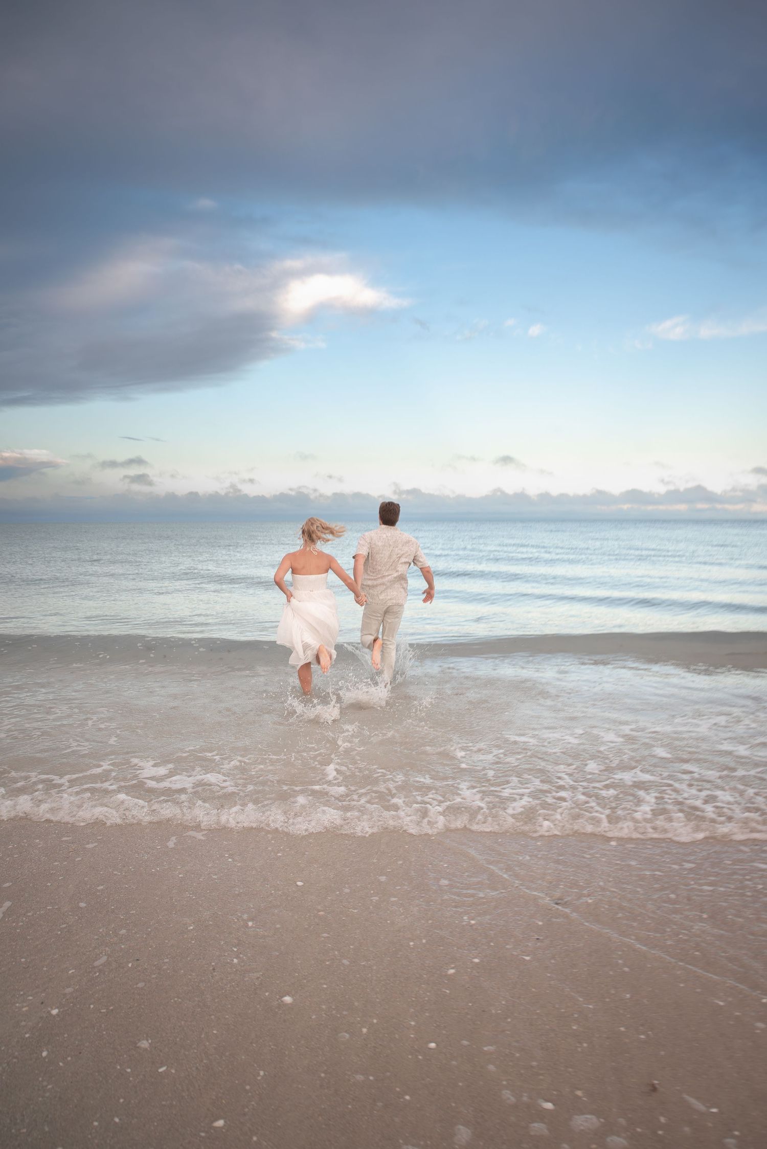 Newlywed couple in wedding attire running hand in hand into ocean waves on sandy beach.
