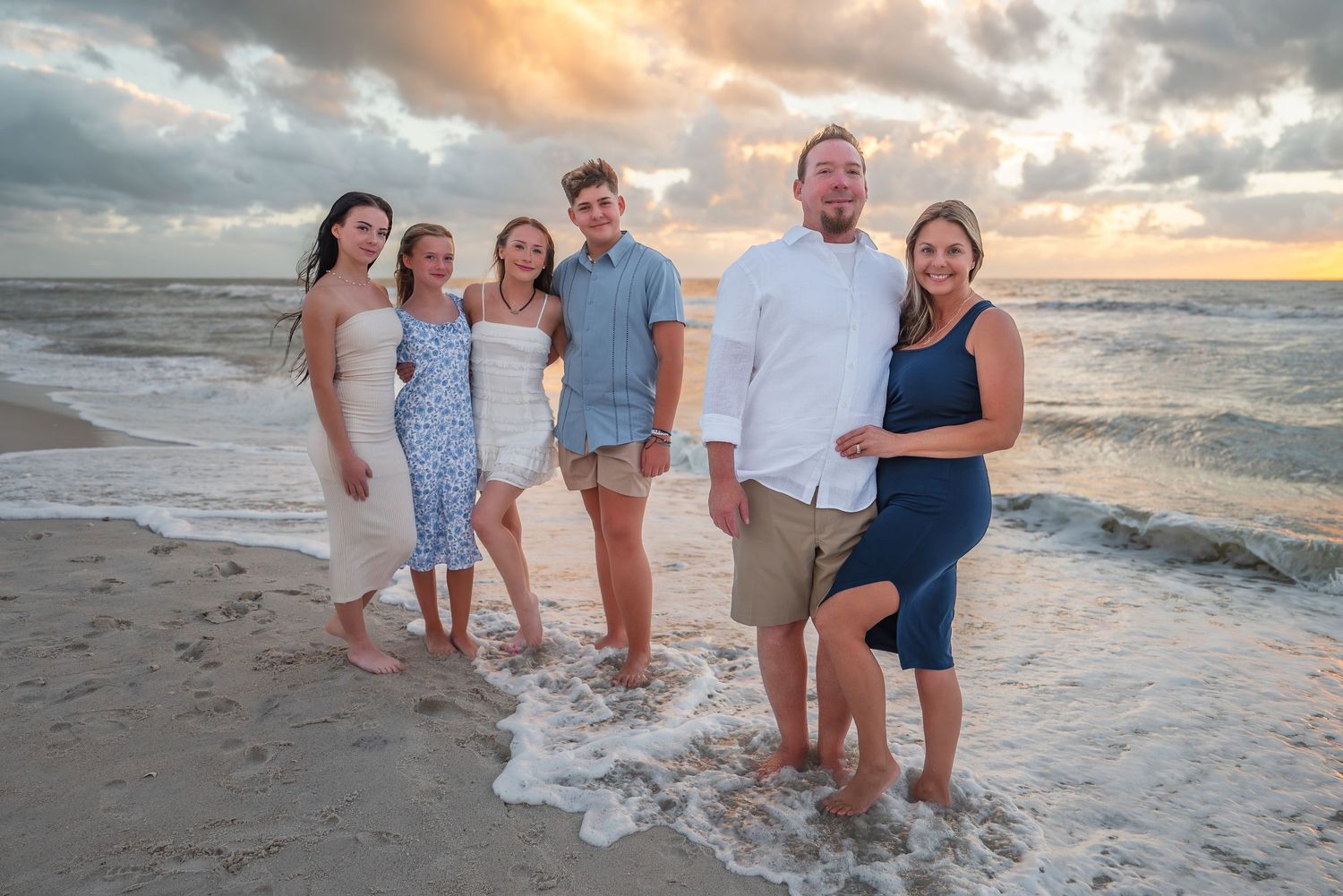 Smiling family of six stands together at the shoreline as waves wash in during a beach sunset.