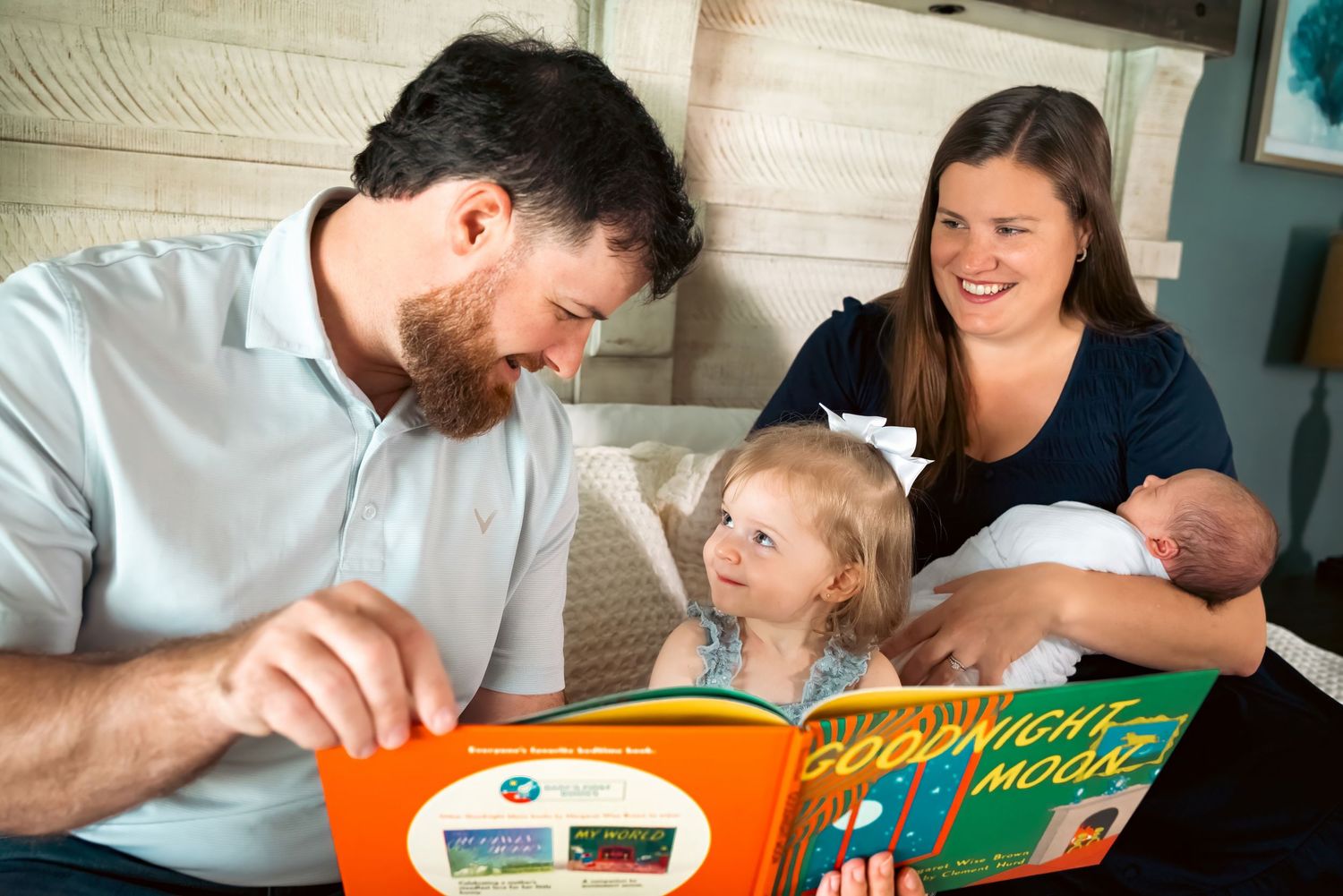 A happy family reads Goodnight Moon together, with parents smiling at their toddler and newborn baby on a cozy bed.
