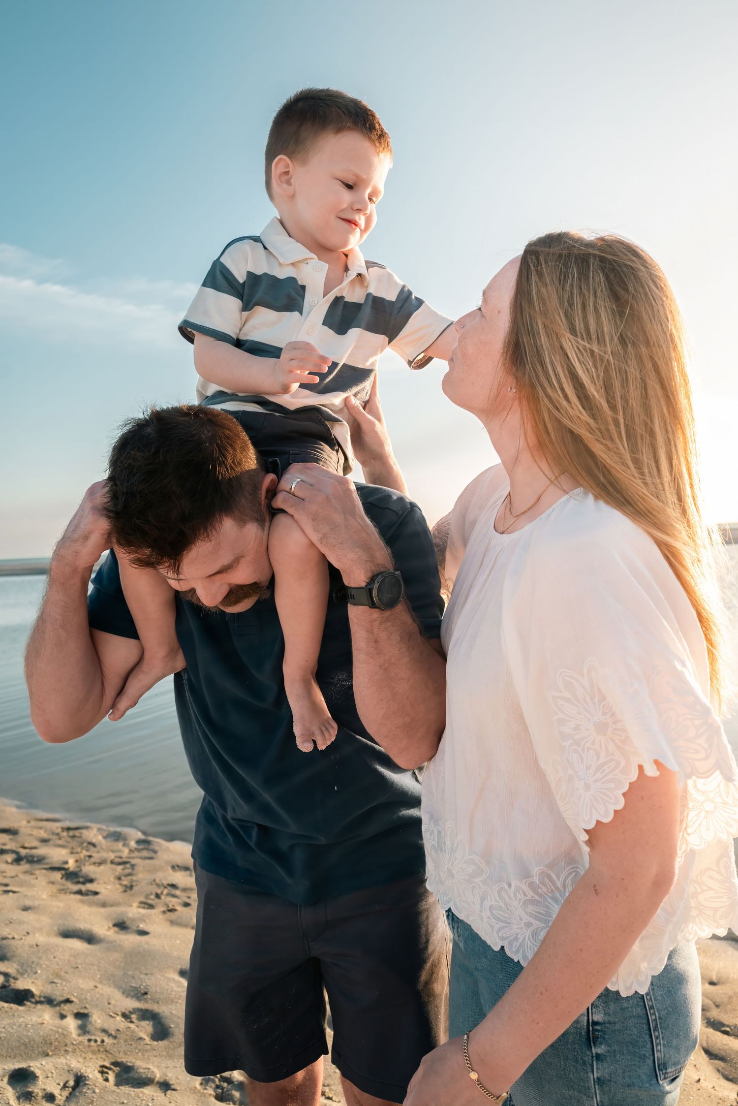 Happy family at the beach, dad carrying toddler on shoulders while mom smiles, golden hour sunset.