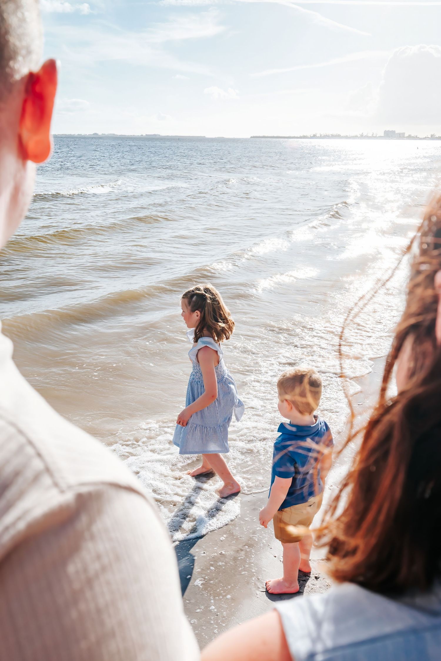 Two children playing in ocean waves on a sunny beach while parents watch from the foreground.