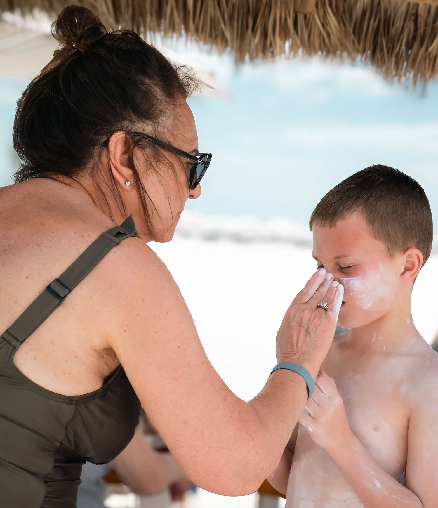 A woman applying sunscreen to a young boy's face on a sunny beach under a thatched umbrella.