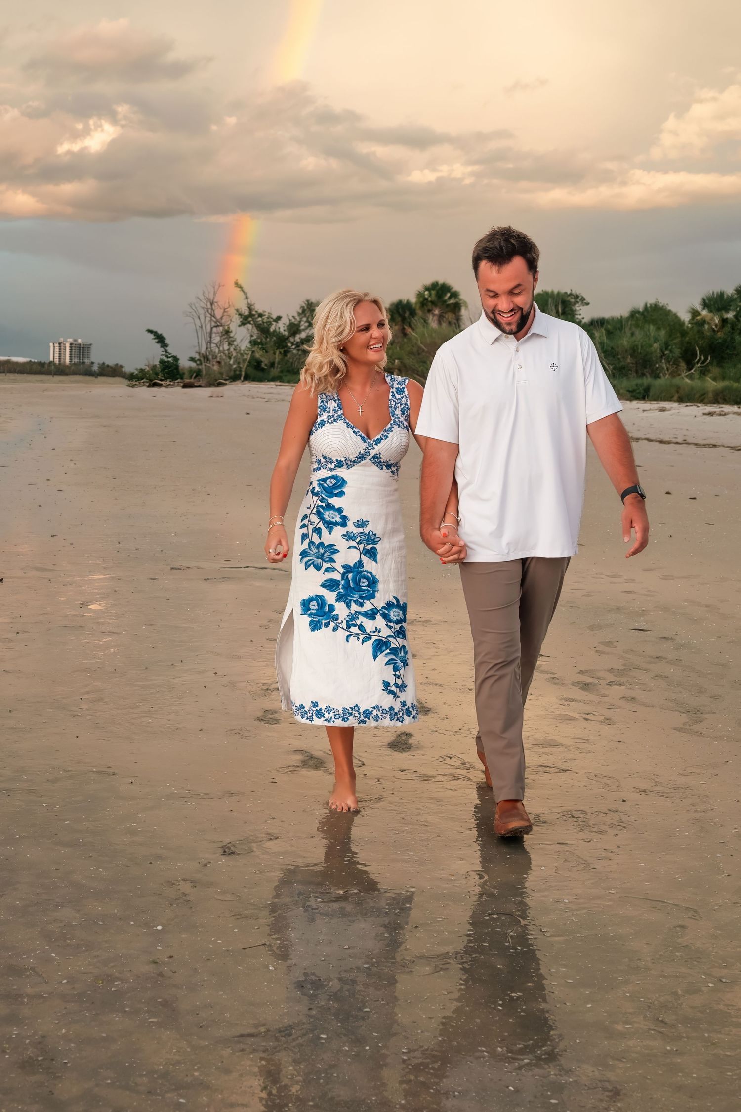 Couple holding hands walking on beach in matching white and blue outfits with rainbow in cloudy sky backdrop.
