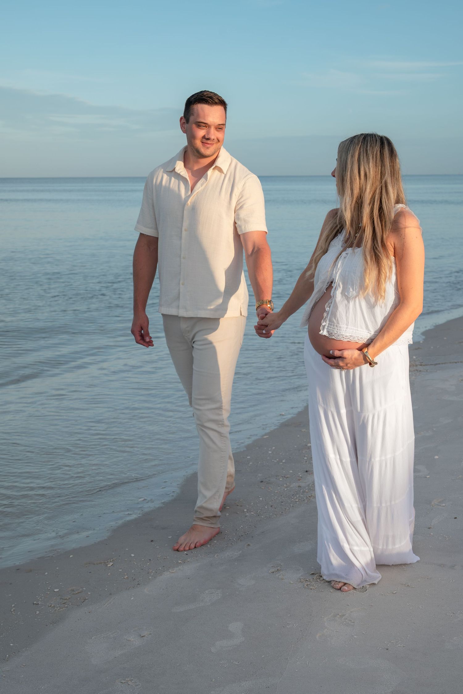 Expecting couple holding hands and walking along the shoreline at sunset, woman in white maternity dress.