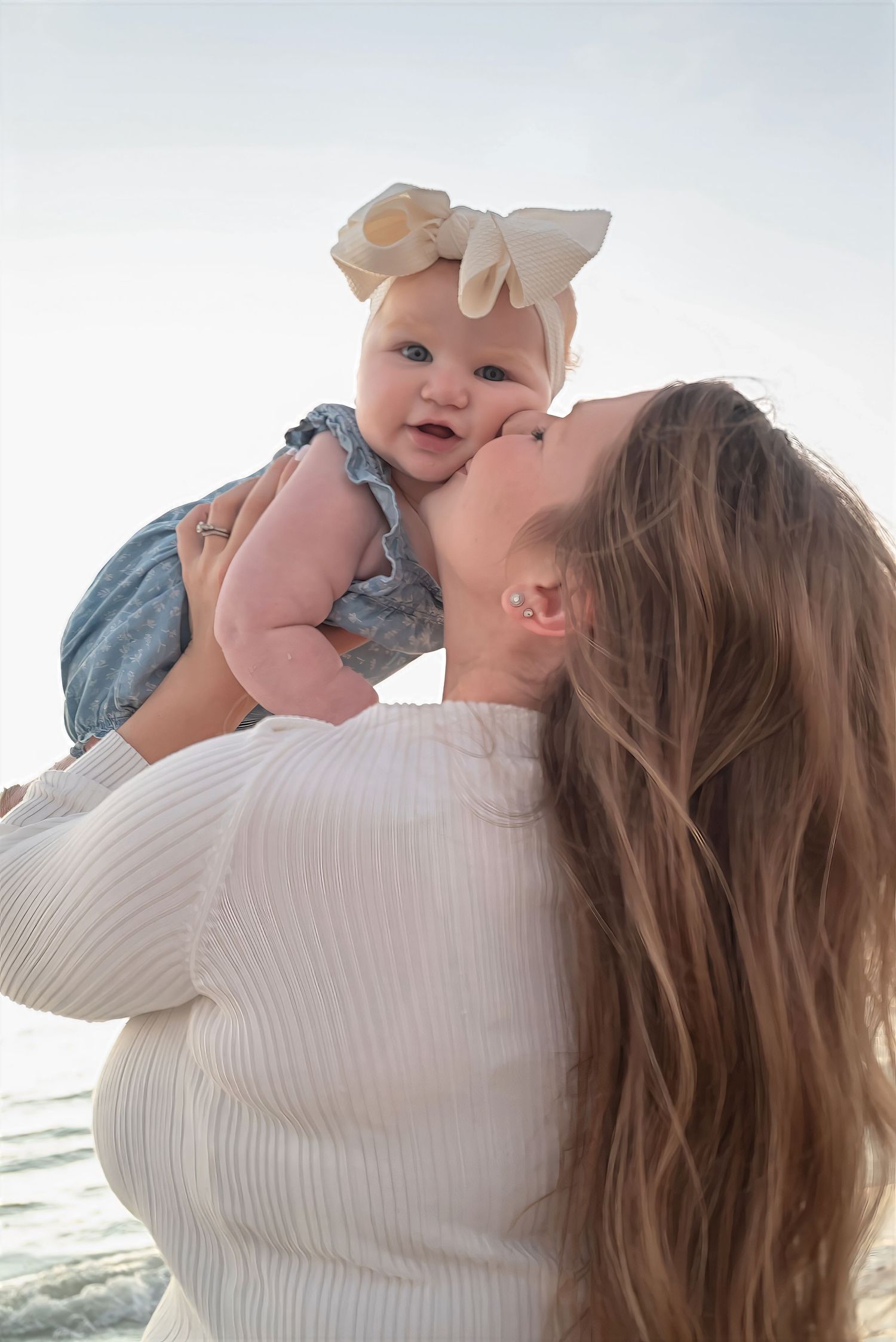 Mother kissing smiling baby girl with bow headband held up against a bright coastal background.