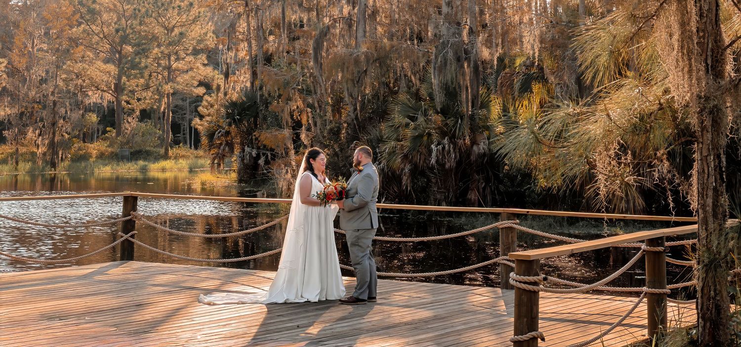 Bride and groom exchange vows on a rustic wooden dock beside a tranquil cypress-lined lake.