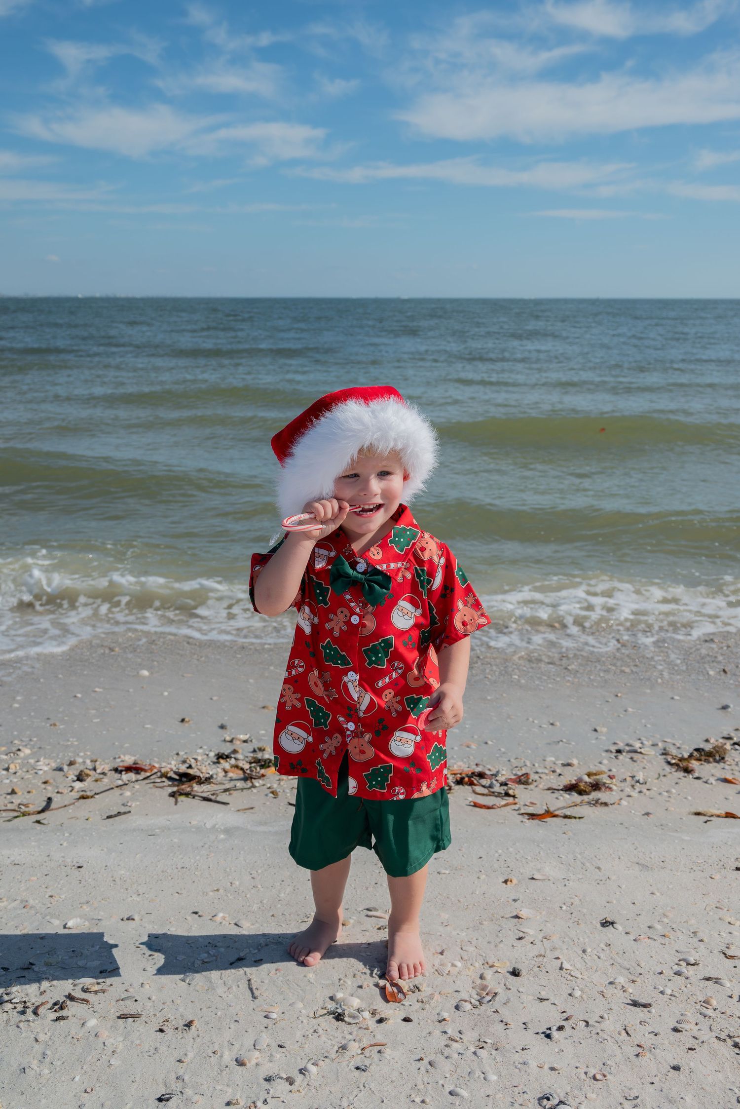 Young boy in Santa hat and Christmas shirt standing barefoot on the beach.