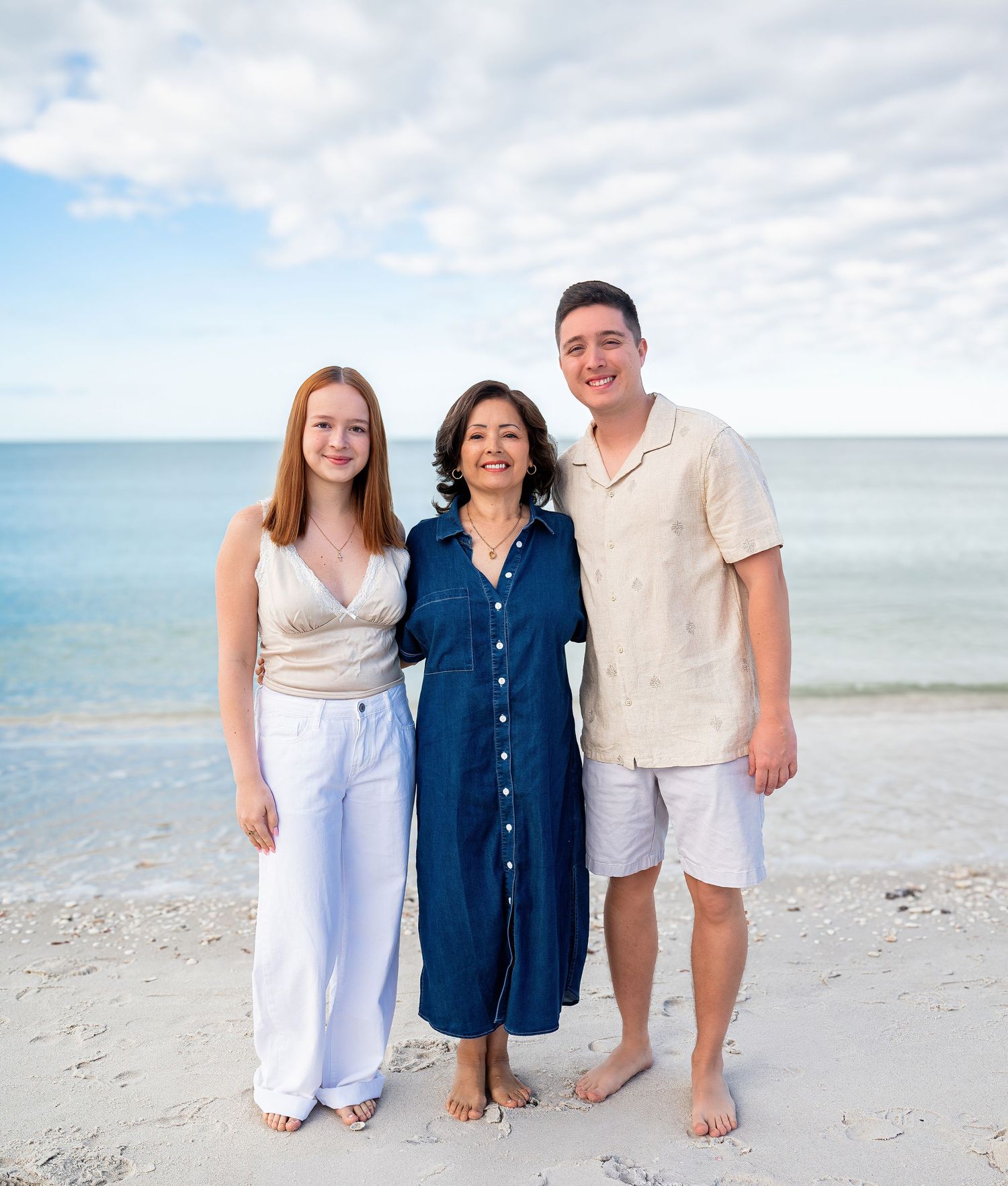 Family of three smiling together on a sunny beach with ocean backdrop.