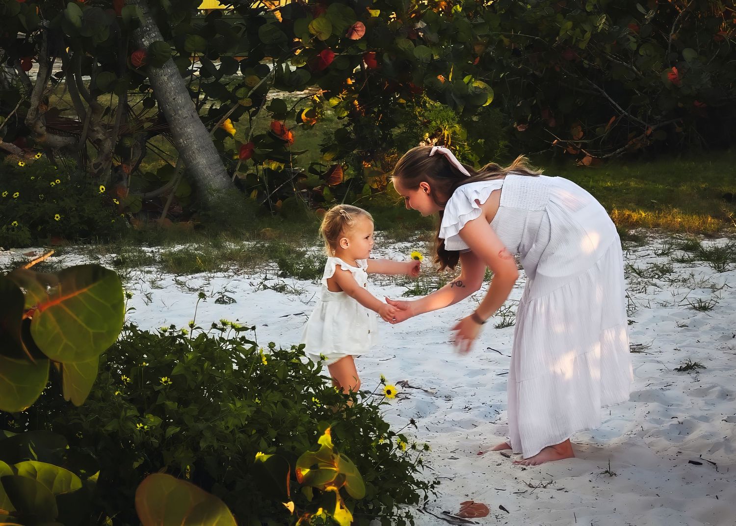 Mother in white dress playfully dancing with toddler daughter on sandy beach among tropical plants.
