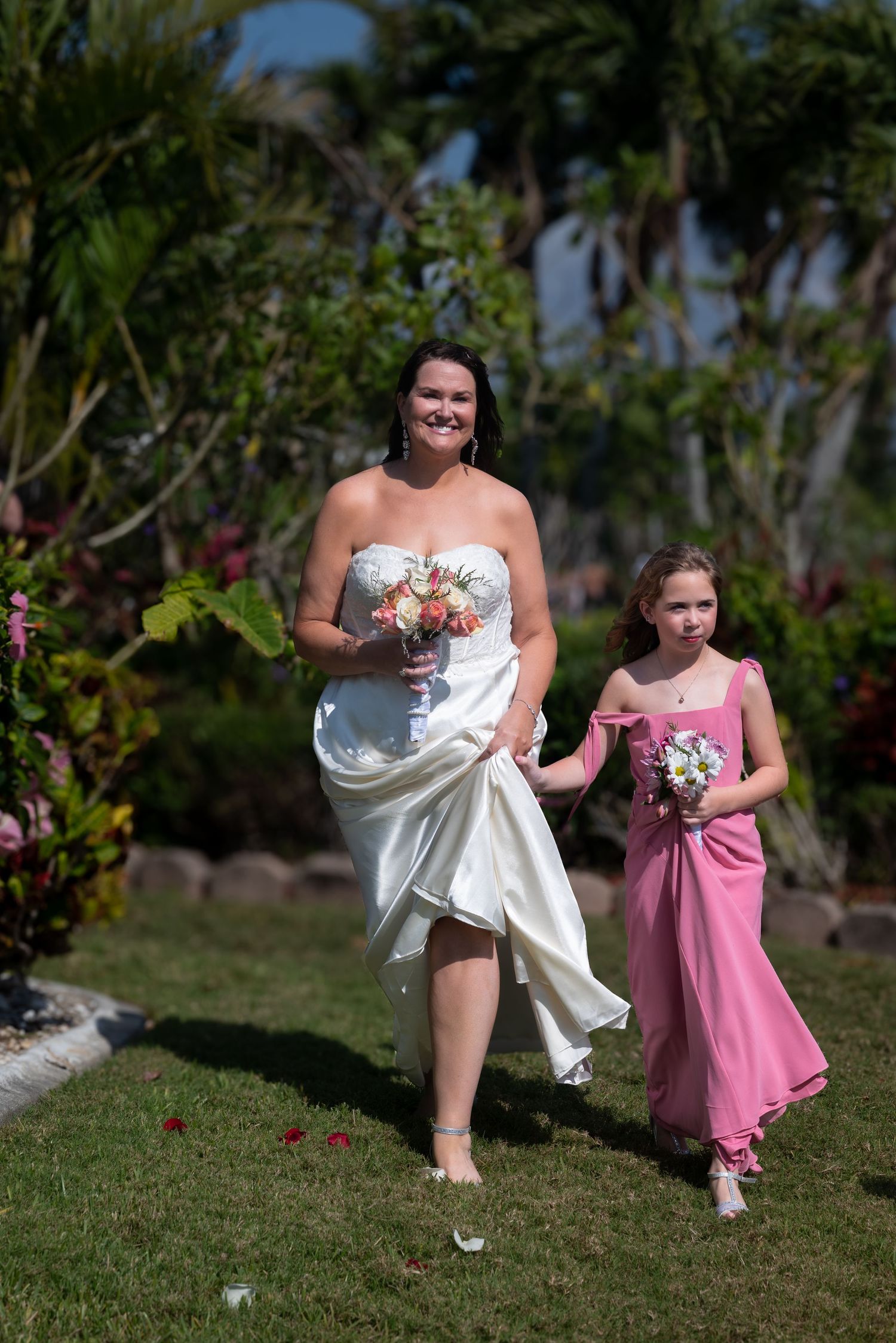 Bride in white strapless gown walks with young flower girl in pink dress carrying bouquets at outdoor wedding.