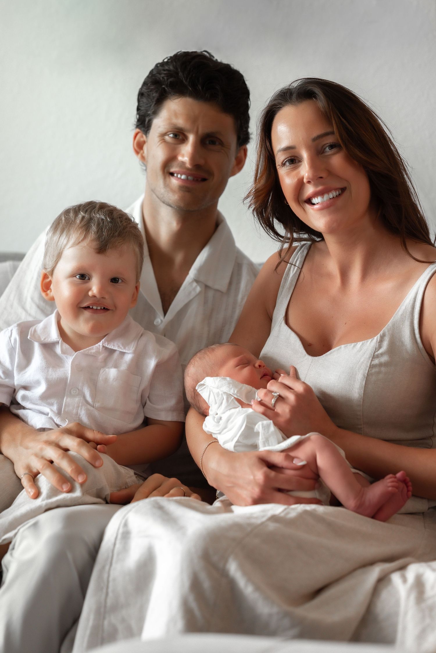 Smiling family portrait with parents, toddler son, and newborn baby sitting together.