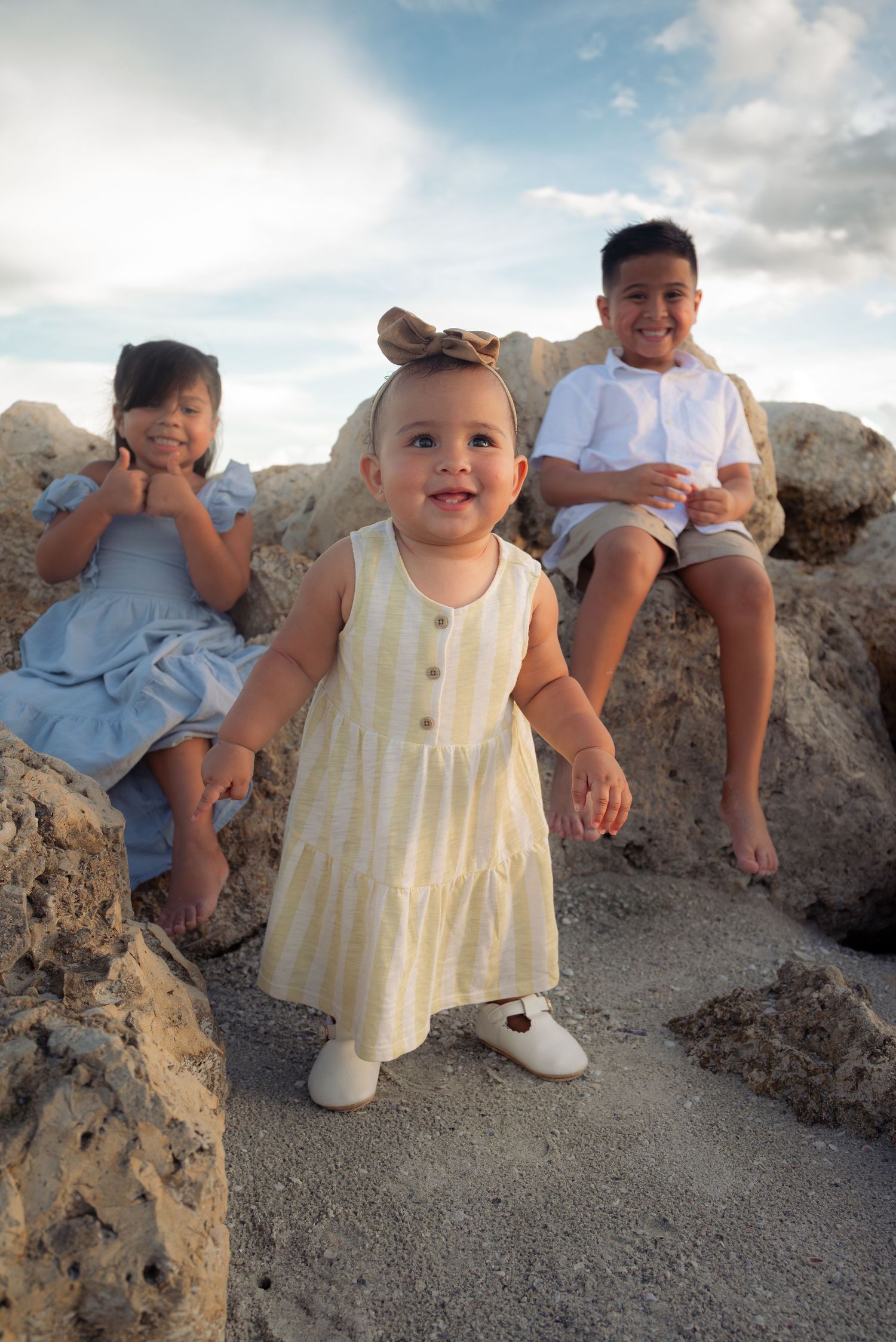 A happy baby girl in a yellow dress stands on rocks at the beach with her older siblings behind her.
