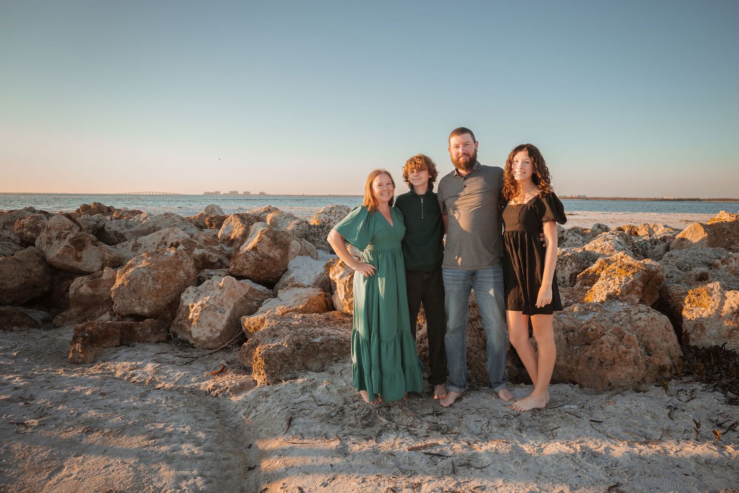 A family of four smiles together on a rocky beach at golden hour sunset.
