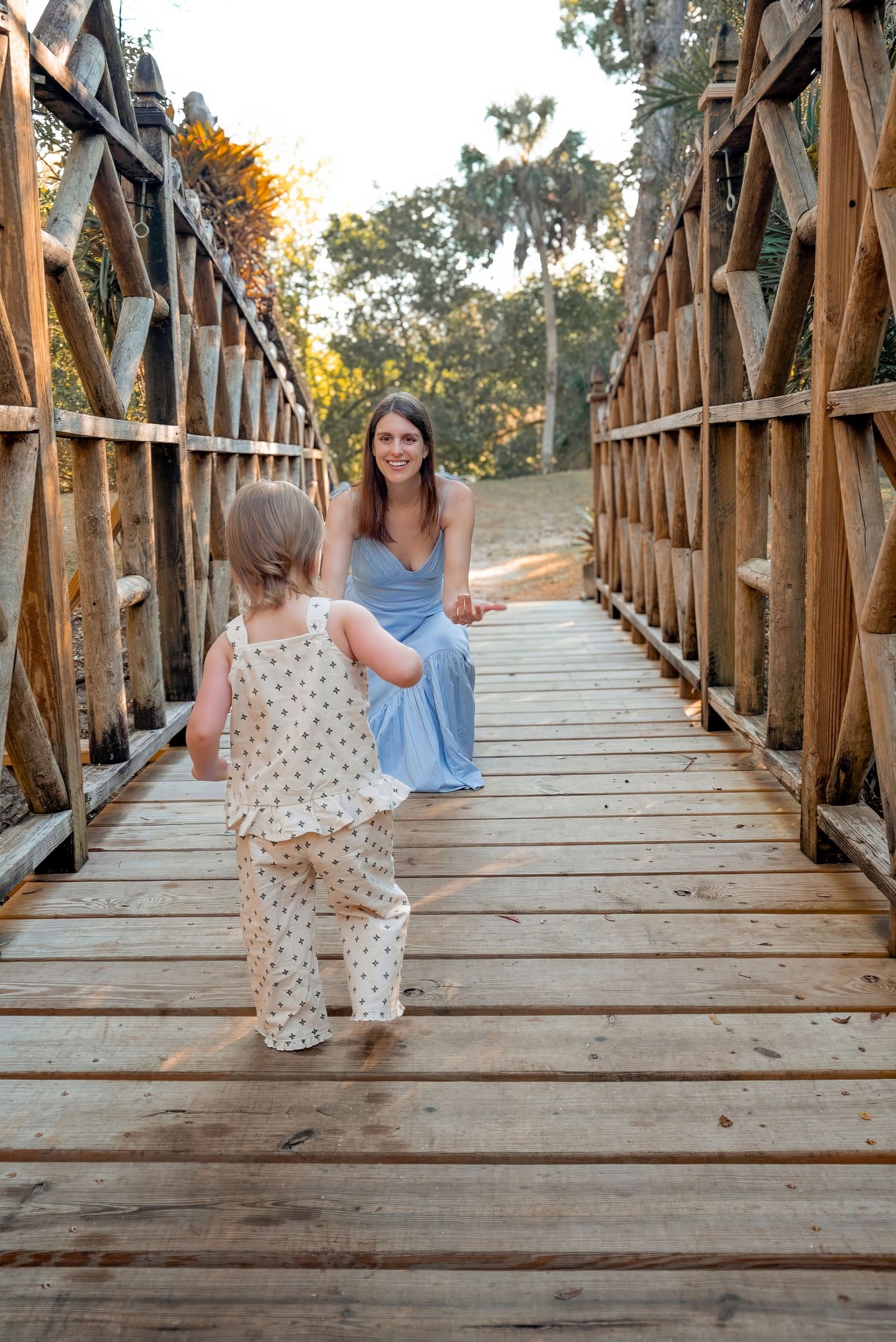 Young child in patterned outfit walking toward smiling mother seated on a rustic wooden bridge.