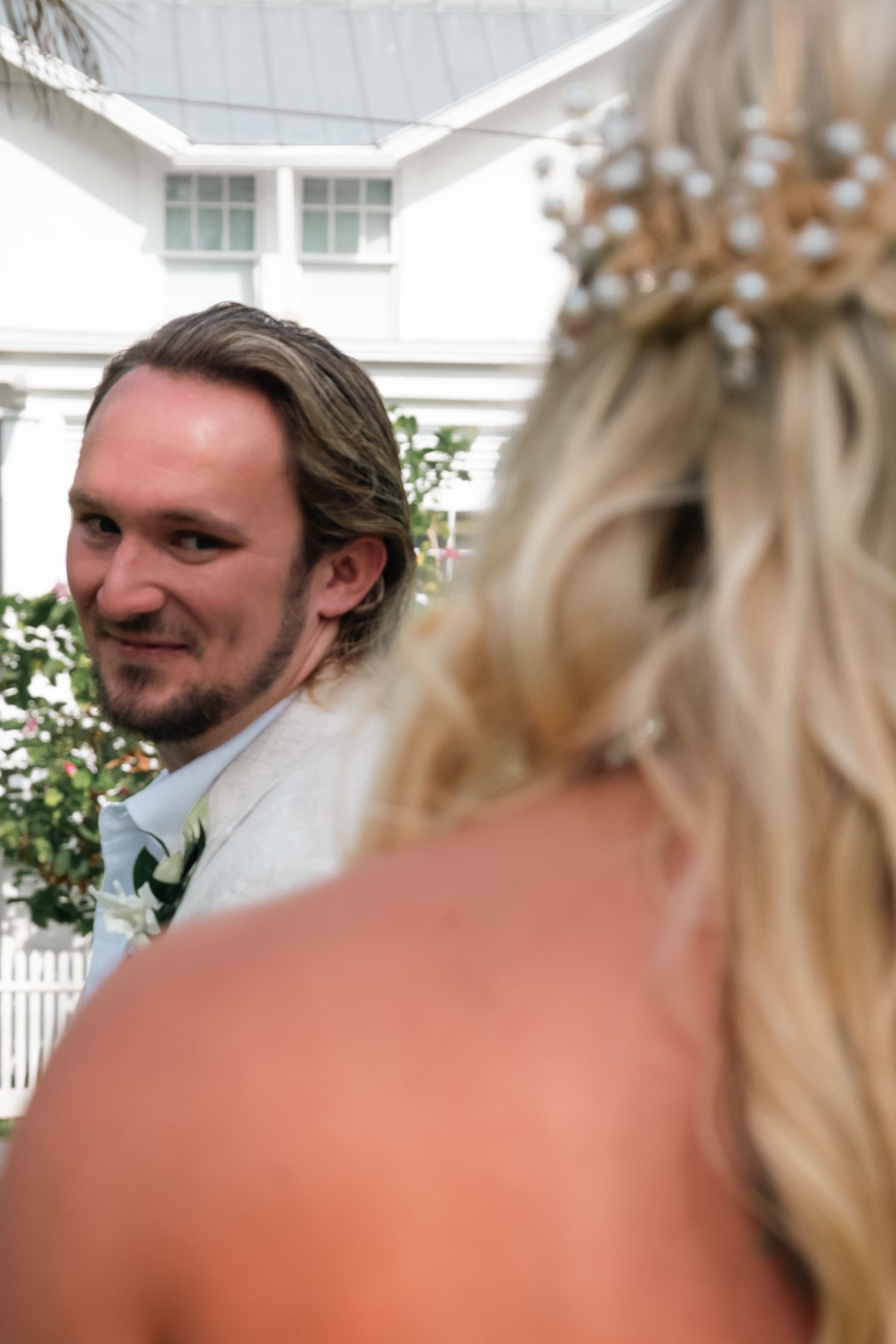 Groom smiling at bride during outdoor wedding ceremony, bride wearing floral crown, white building in background.