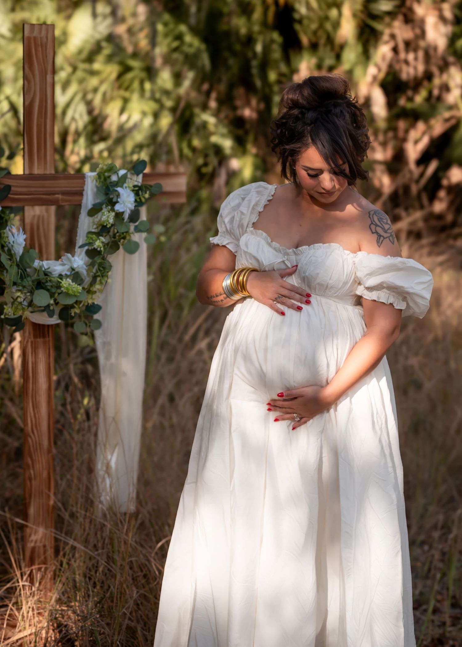 Pregnant woman in white off-shoulder gown cradling belly near wooden cross with floral wreath outdoors.
