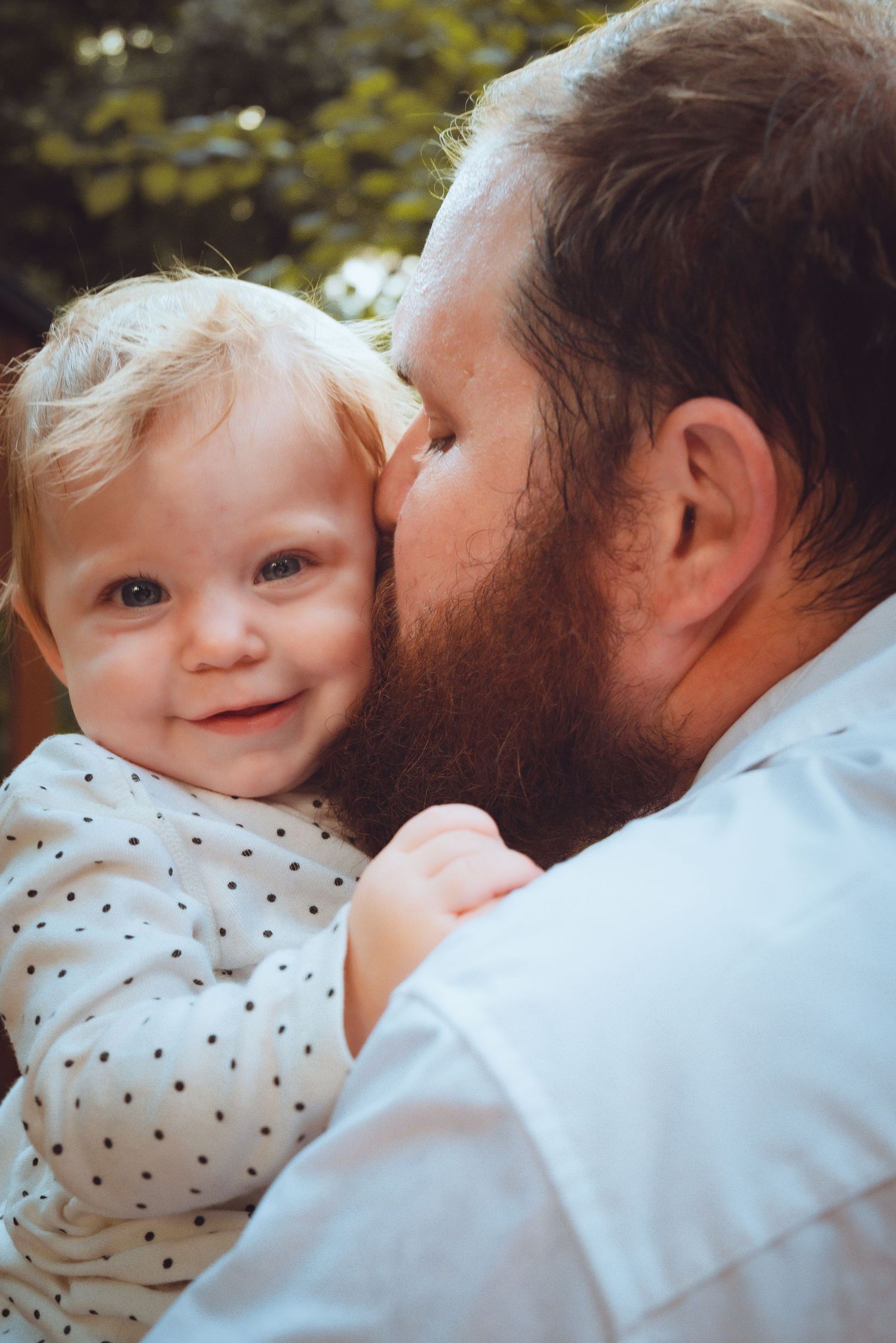 A bearded father gently kisses his smiling blonde toddler on the cheek outdoors.