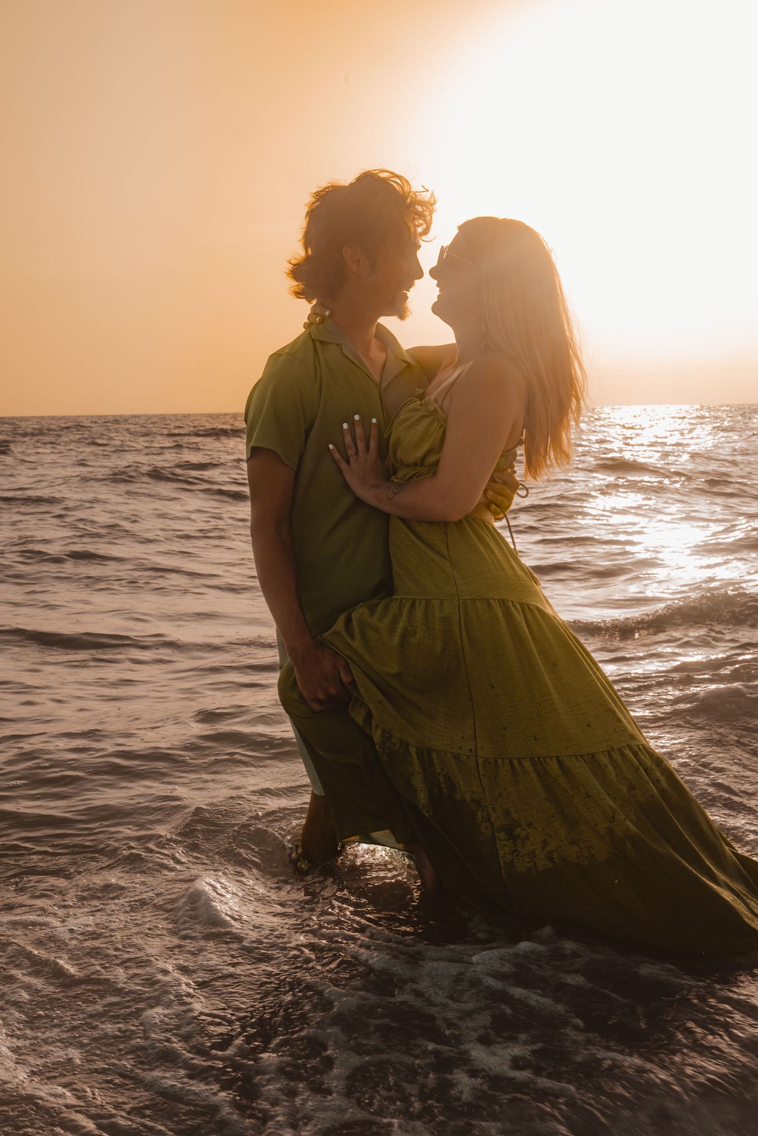 Romantic couple embracing and kissing in ocean waves at sunset, both wearing green outfits in golden light.