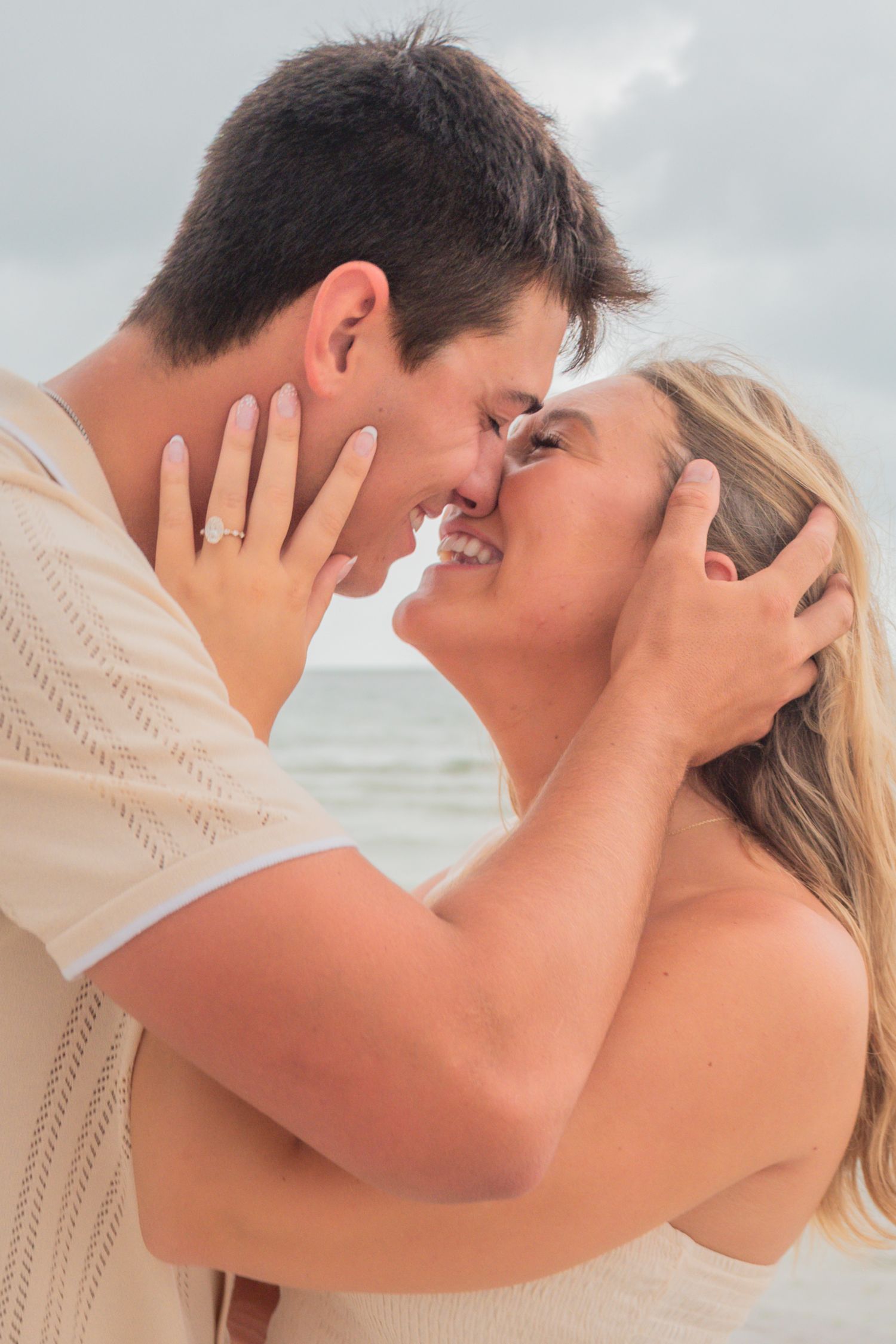 Couple sharing a romantic almost-kiss on the beach, man holding woman's face with engagement ring visible.