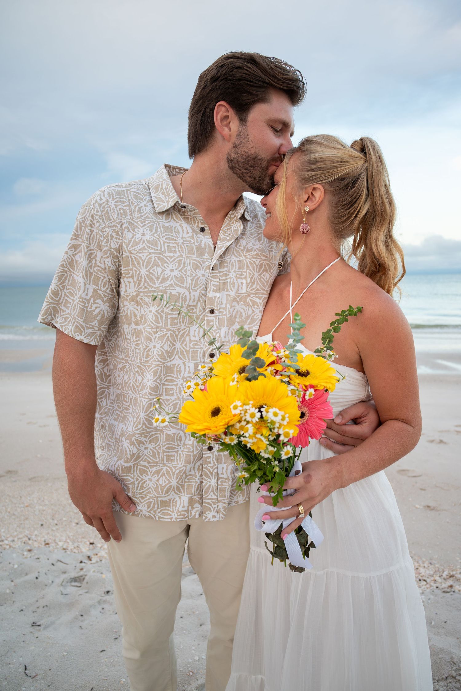 Couple almost kissing on sandy beach, bride holding vibrant yellow gerbera daisy bouquet in white sundress.
