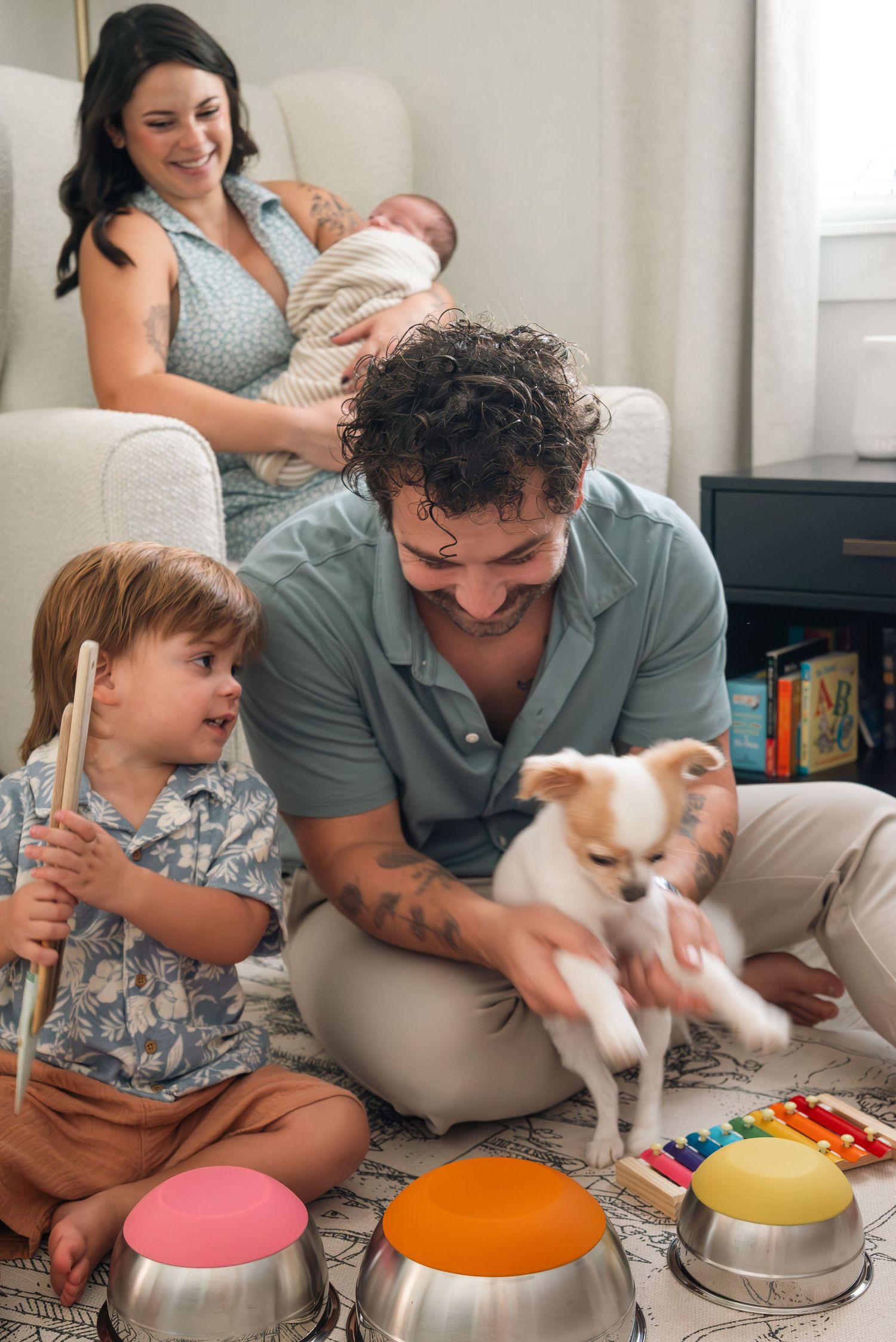 Happy family with newborn baby, toddler playing drums, dad holding small dog in cozy living room.